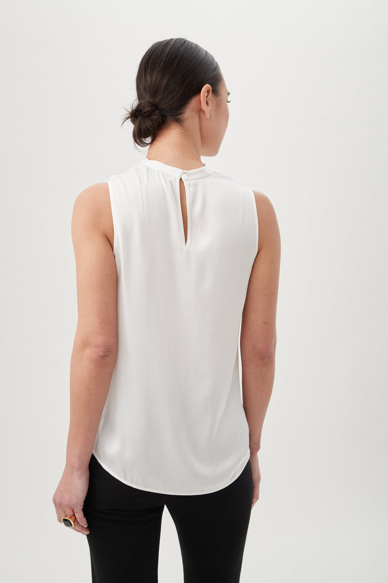A woman with dark hair in a low bun is seen from behind wearing the Lenaya Top, a sleeveless stretch satin style with a small back keyhole, paired with black pants against a plain white background.