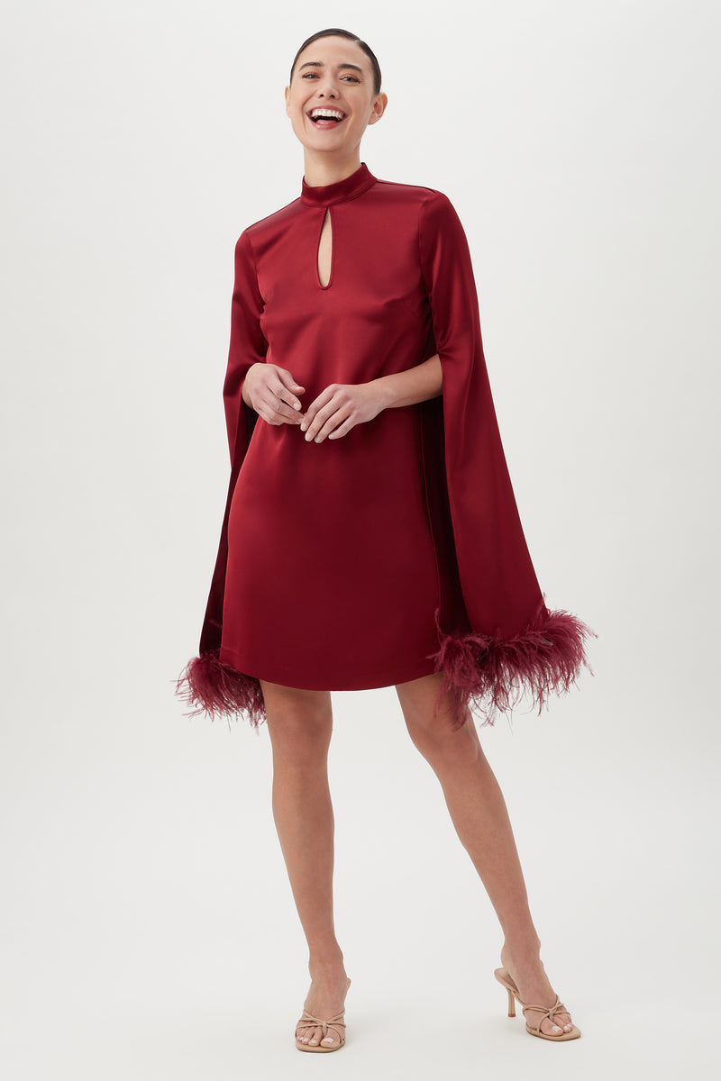 A woman smiles in the Hiromi 2 Dress in Ruqa Red, featuring a short satin mockneck design with a keyhole cutout, cape sleeves, and feather-trimmed cuffs. She stands in open-toe heels against a plain white background.
