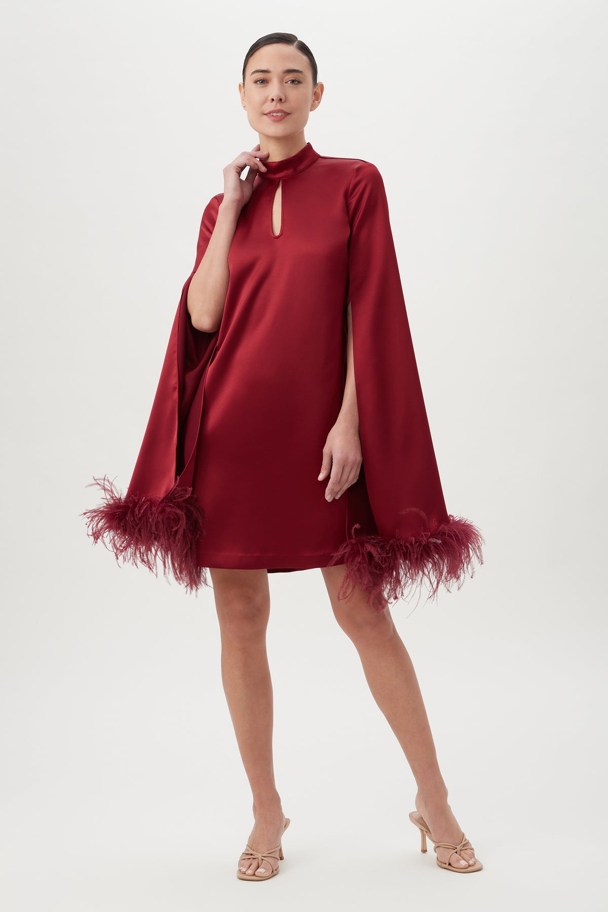 A woman poses confidently in the Hiromi 2 DressÃ¢â‚¬â€a knee-length, long-sleeved burgundy satin mockneck dress with feather-trimmed cuffs, sheath silhouette, and front keyhole cutoutÃ¢â‚¬â€paired with open-toe heels against a plain background.