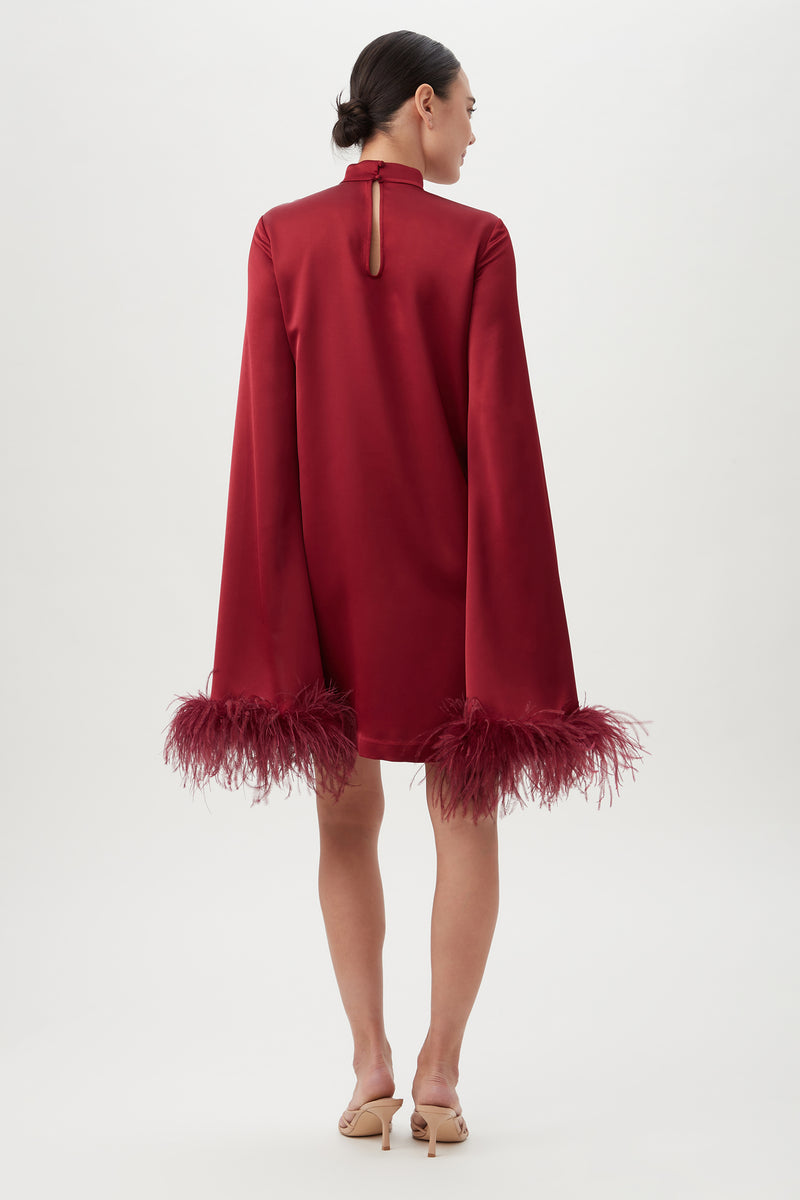 A woman with a low bun faces away, wearing the Hiromi 2 DressÃ¢â‚¬â€a red satin mockneck style with long, wide sleeves and feathery cuff trim. She completes the look with nude heeled sandals against a plain, light background.