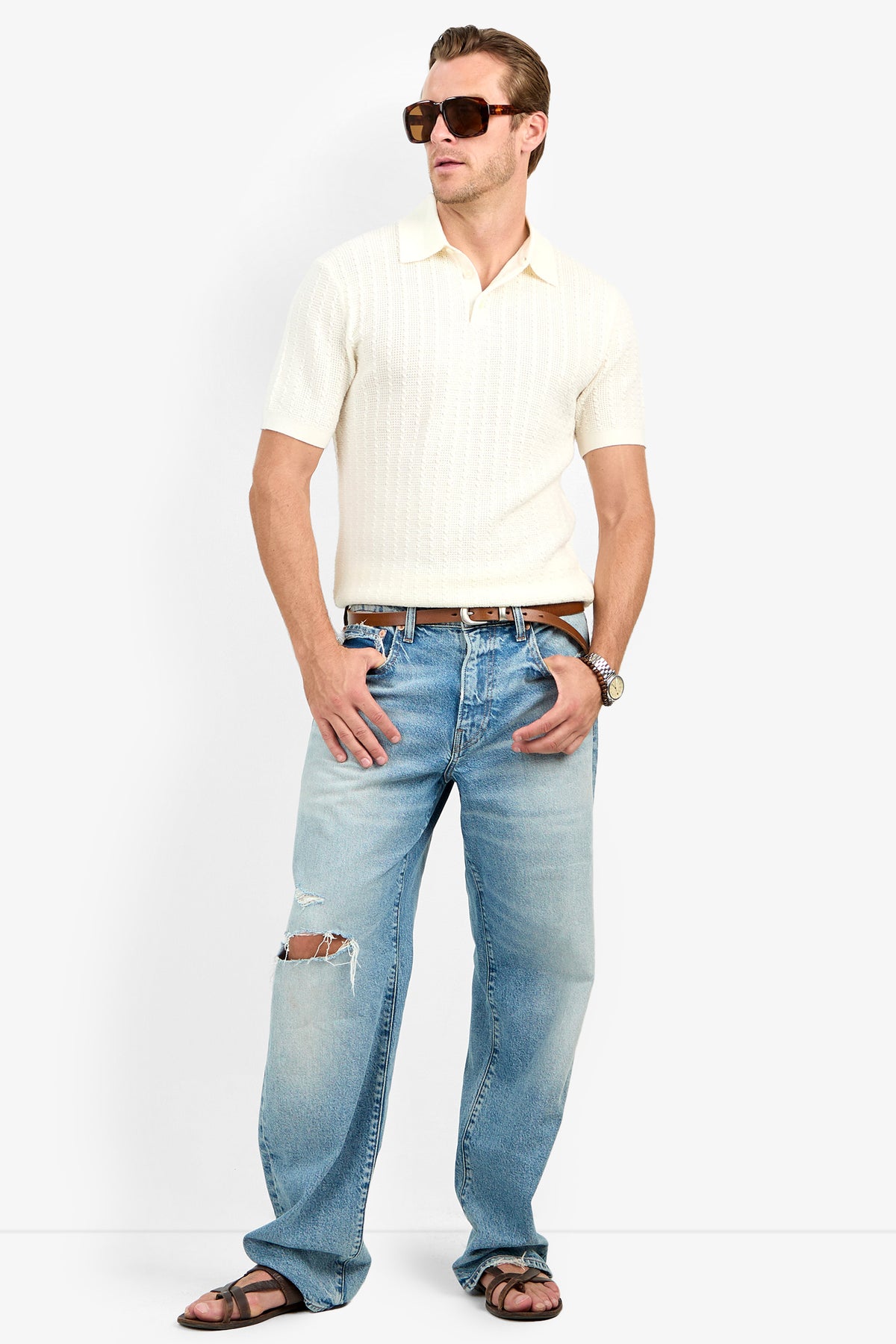 Man in large sunglasses, Asher Polo in white, blue distressed men’s slim fit jeans with a knee rip, brown sandals, and a watch stands with one hand in his pocket against a plain background.