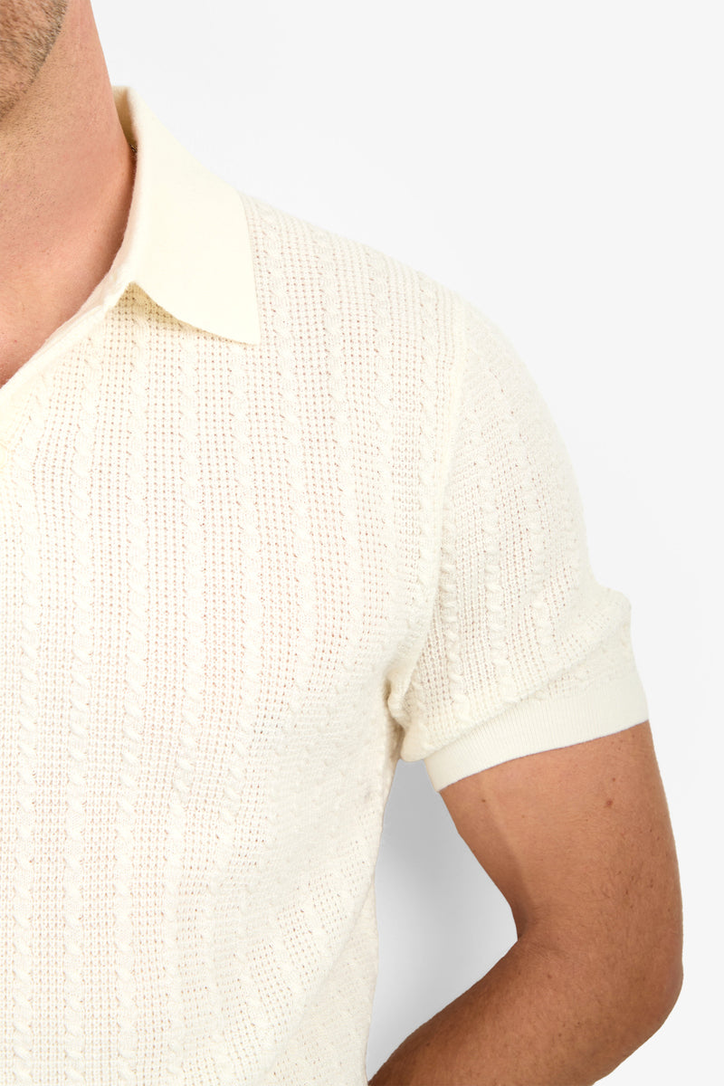A close-up of a person wearing the cream-colored Asher Polo, featuring short sleeves, cable knit Merino wool, and a collar. Only the upper torso and part of one arm and shoulder are visible.