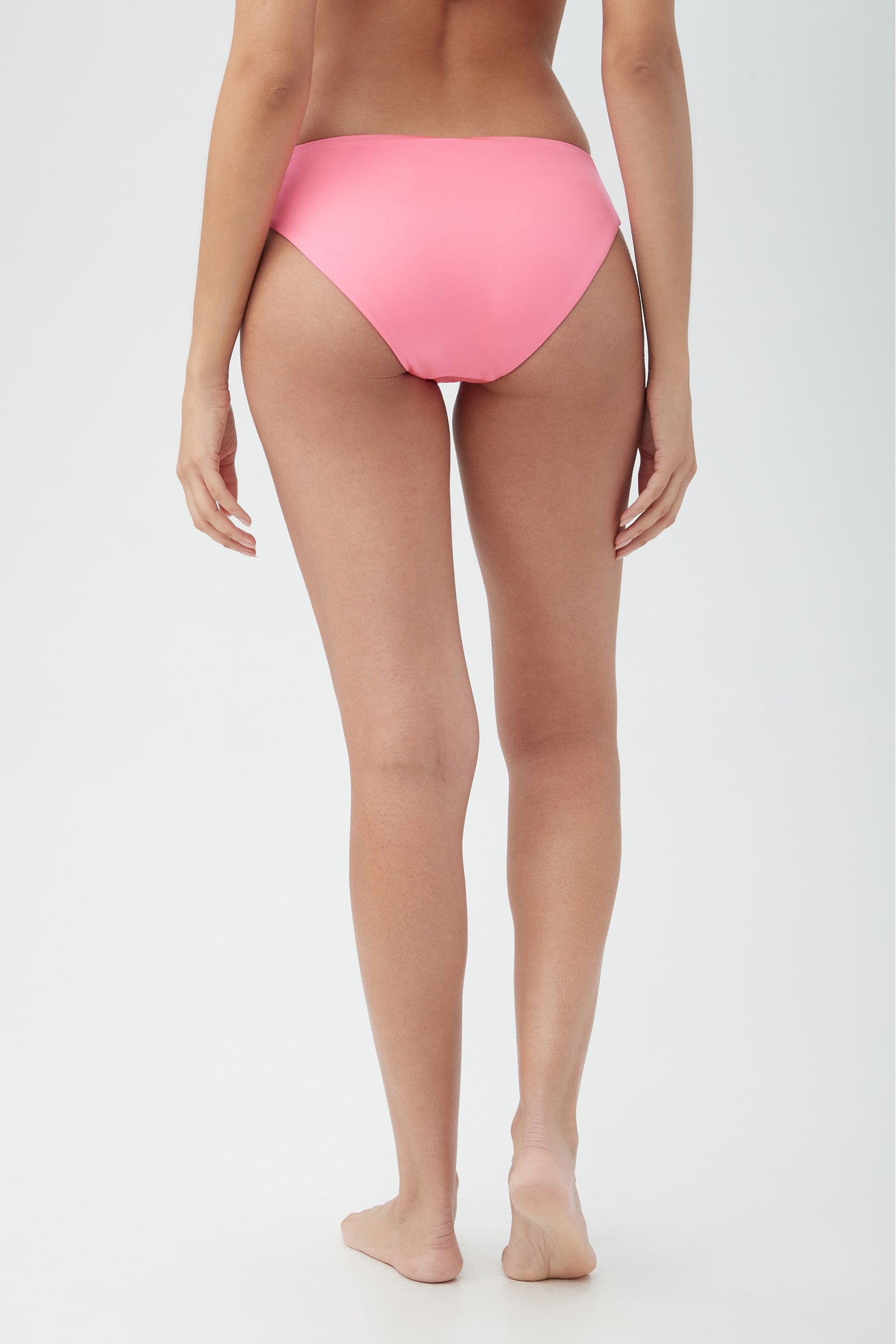 A woman is standing barefoot with her back to the camera, wearing the Women's Monaco Shirred Hipster Swim Bottom in pink against a plain light background.