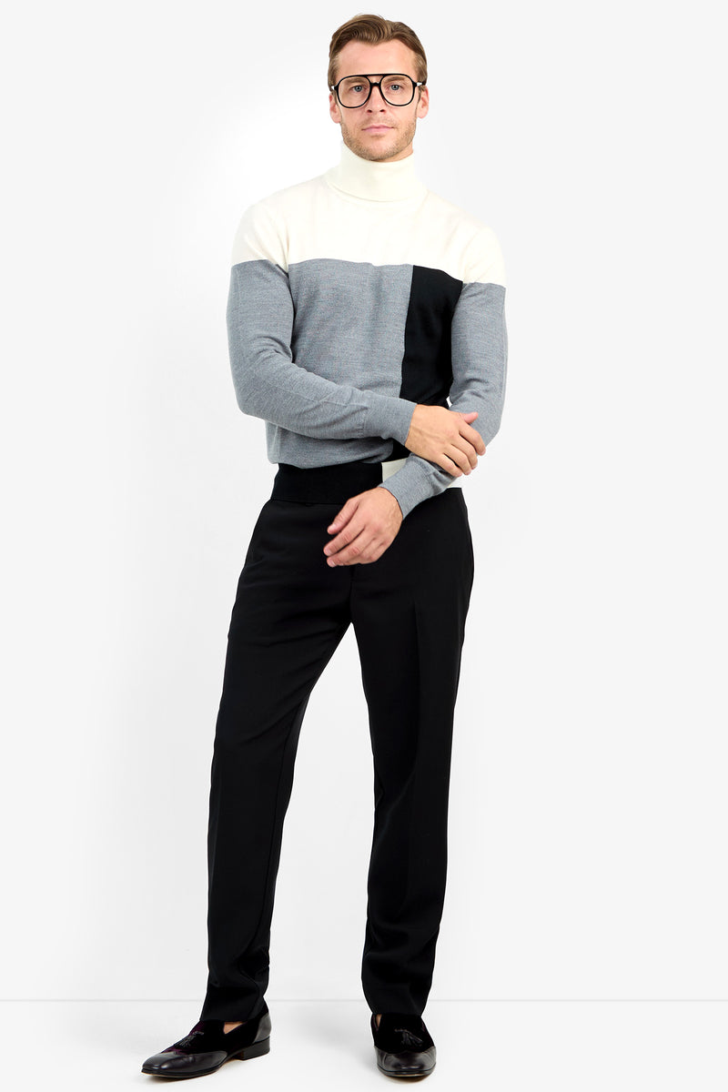 A man in glasses wears the Callisto Sweater—white, gray, and black color-blocked—paired with black pants, standing with arms lightly crossed against a plain white background.