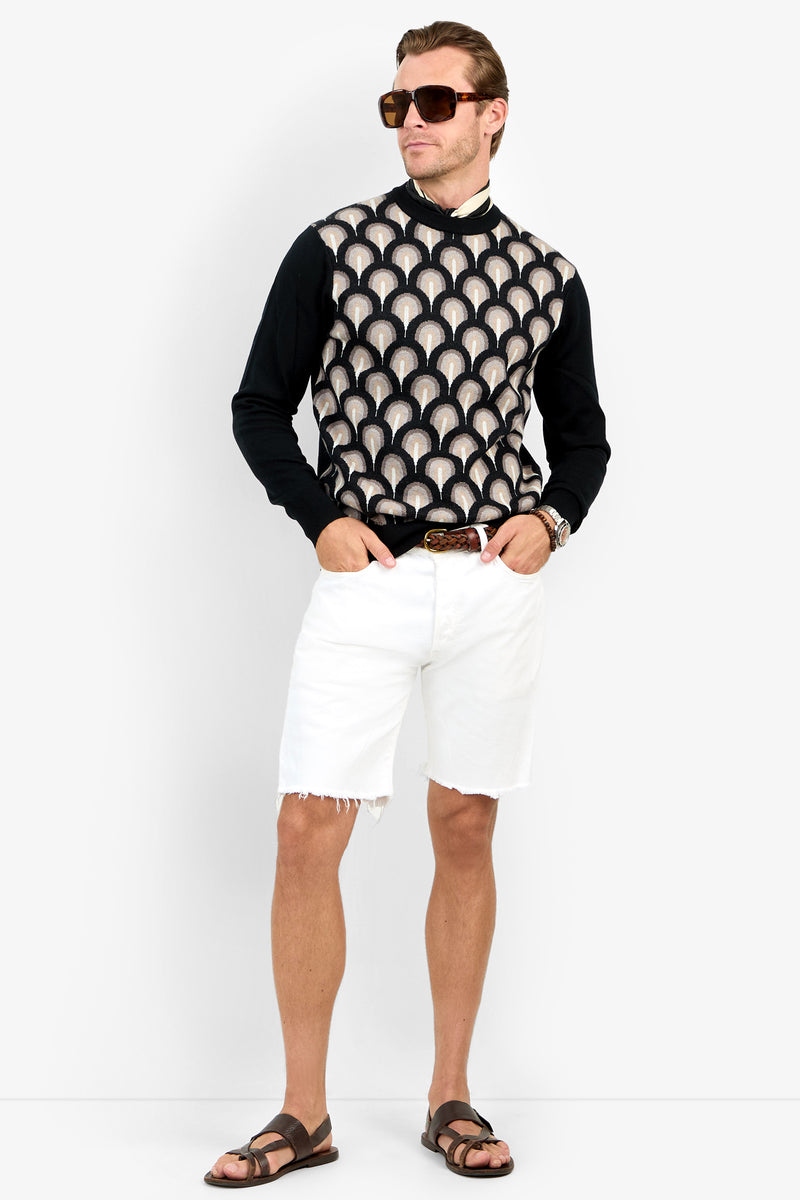A man in sunglasses, the Chrysler Sweater over a black shirt, white frayed shorts, and brown sandals stands against a plain white background with one hand in his pocket.
