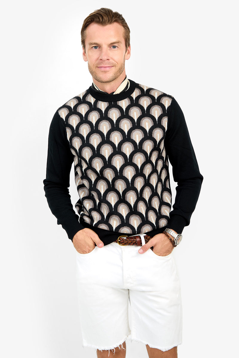 A man wearing the Chrysler Sweater, featuring a black base with beige and white geometric patterns, and white shorts stands smiling with his hands in his pockets.
