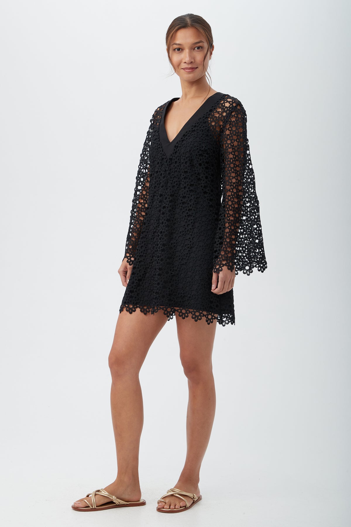 A woman stands against a plain background, smiling slightly with her hair tied back, wearing the Women's Chateau Lace Long Sleeve V-Neck Swim Cover-Up Dress in black and beige flat sandals.