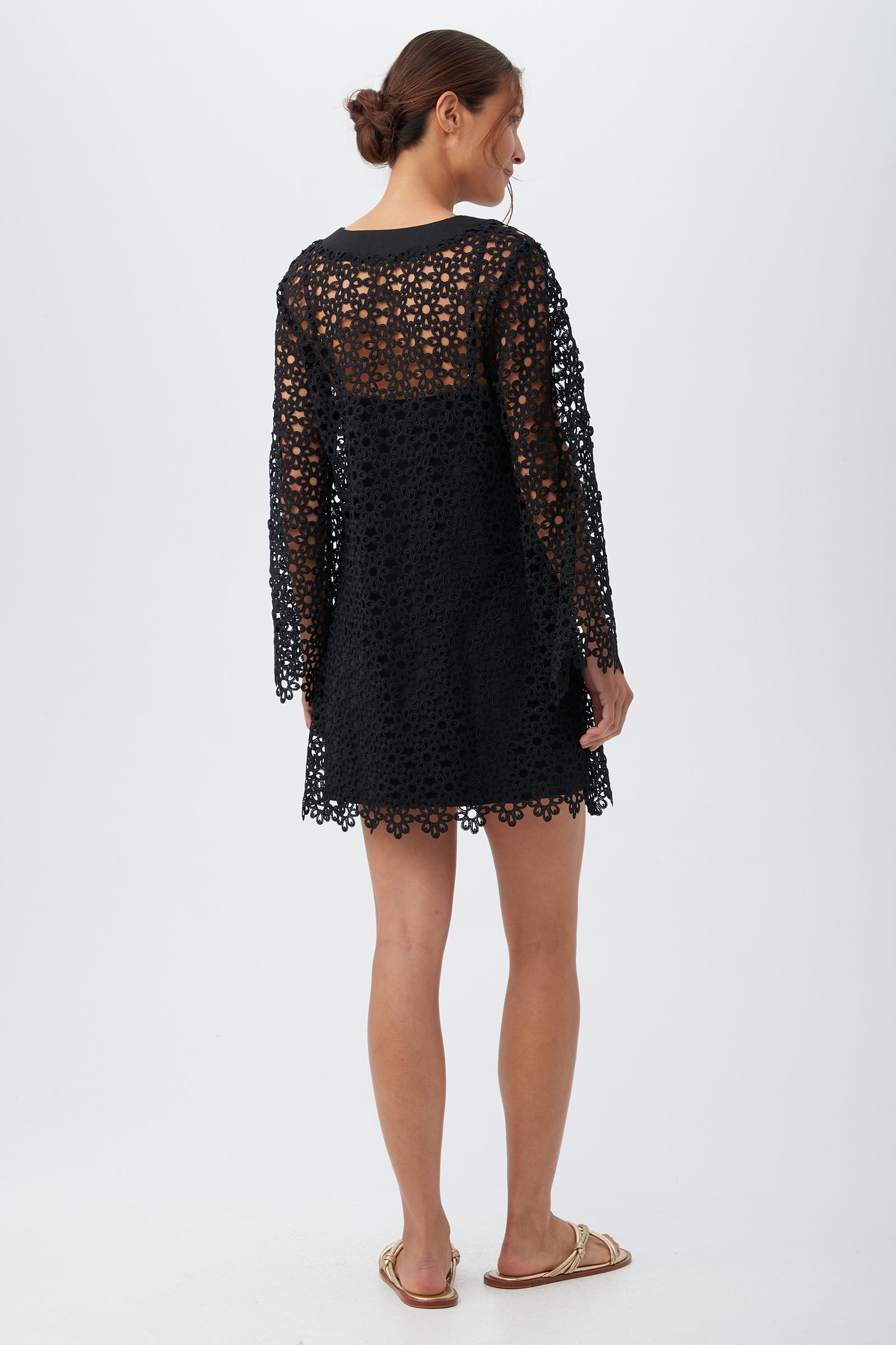 A woman stands facing away, wearing the Women's Chateau Lace Long Sleeve V-Neck Swim Cover-Up Dress in black. Her hair is tied back, and she's wearing flat gold sandals against a plain, light background.