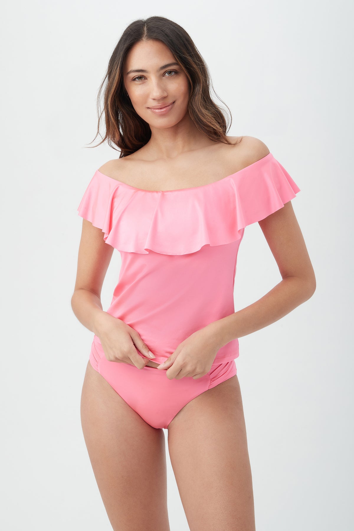 A woman smiles softly with her hands near her waist, wearing the Women's Monaco Off The Shoulder Ruffle Tankini Swim Top in Carnation Pink against a plain light background.