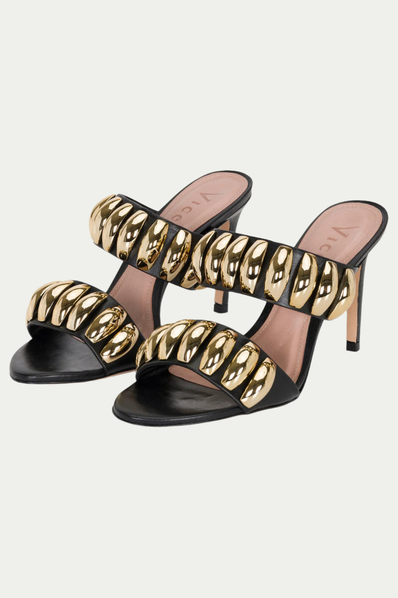 The GOLDEN VOLUME HEEL SANDAL is a black and gold pair of stilettos with two wide straps, each decorated with shiny gold oval embellishments.