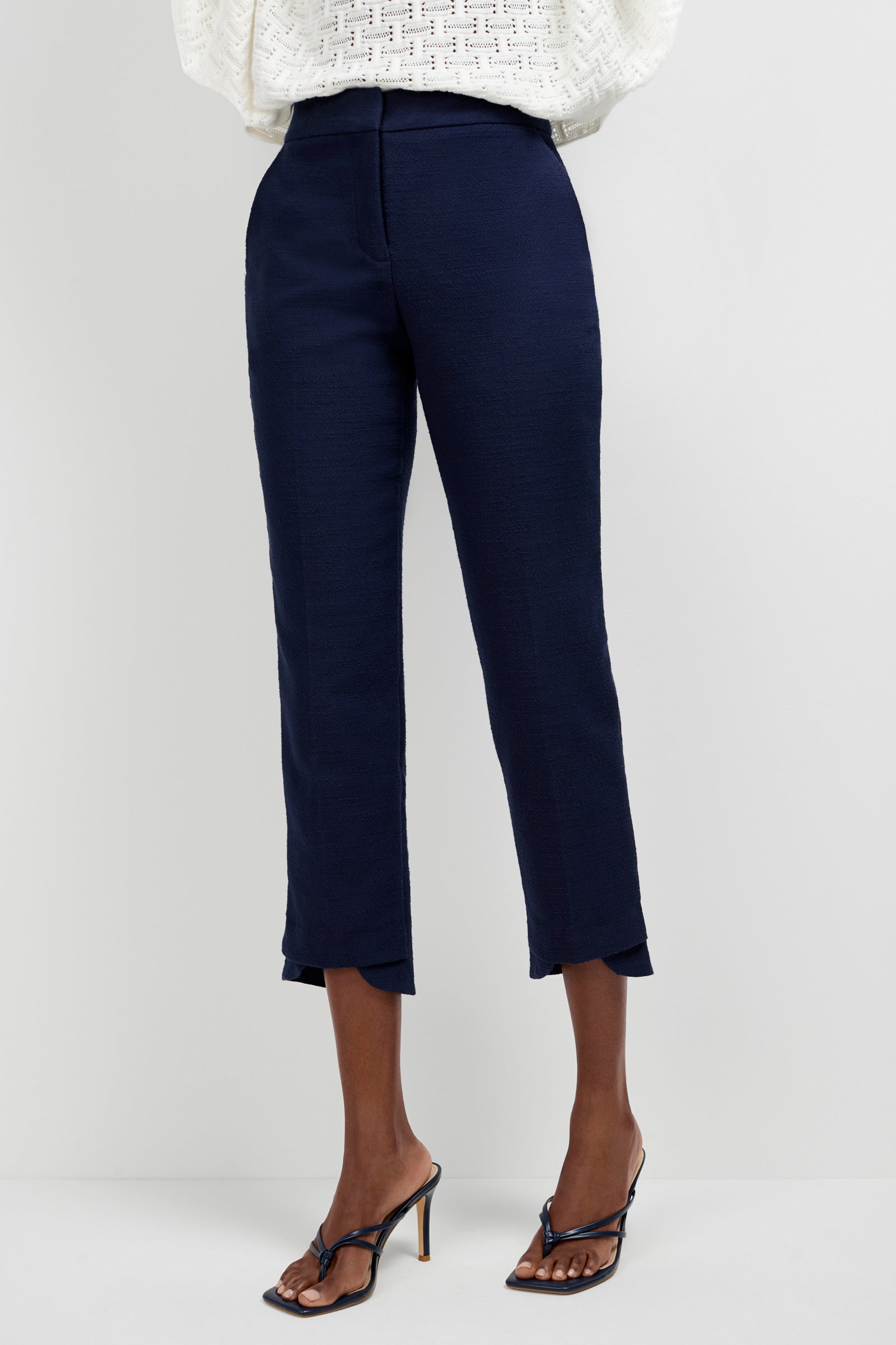 A person stands against a plain light background, showcasing the lower half of their outfit featuring the Ore Button-Hem Cropped Pants in navy, paired with a white textured long-sleeve top and black high-heeled sandals.