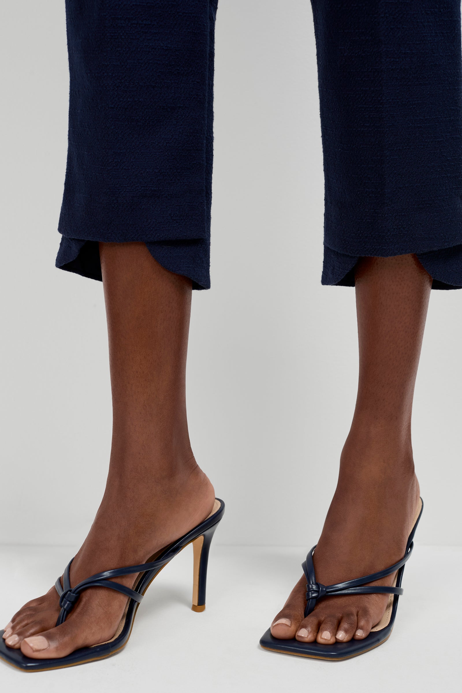 A person wears Ore Button-Hem Cropped Pants in navy with dark, high-heeled, square-toe, open-toe sandals featuring thin straps, standing against a light background.
