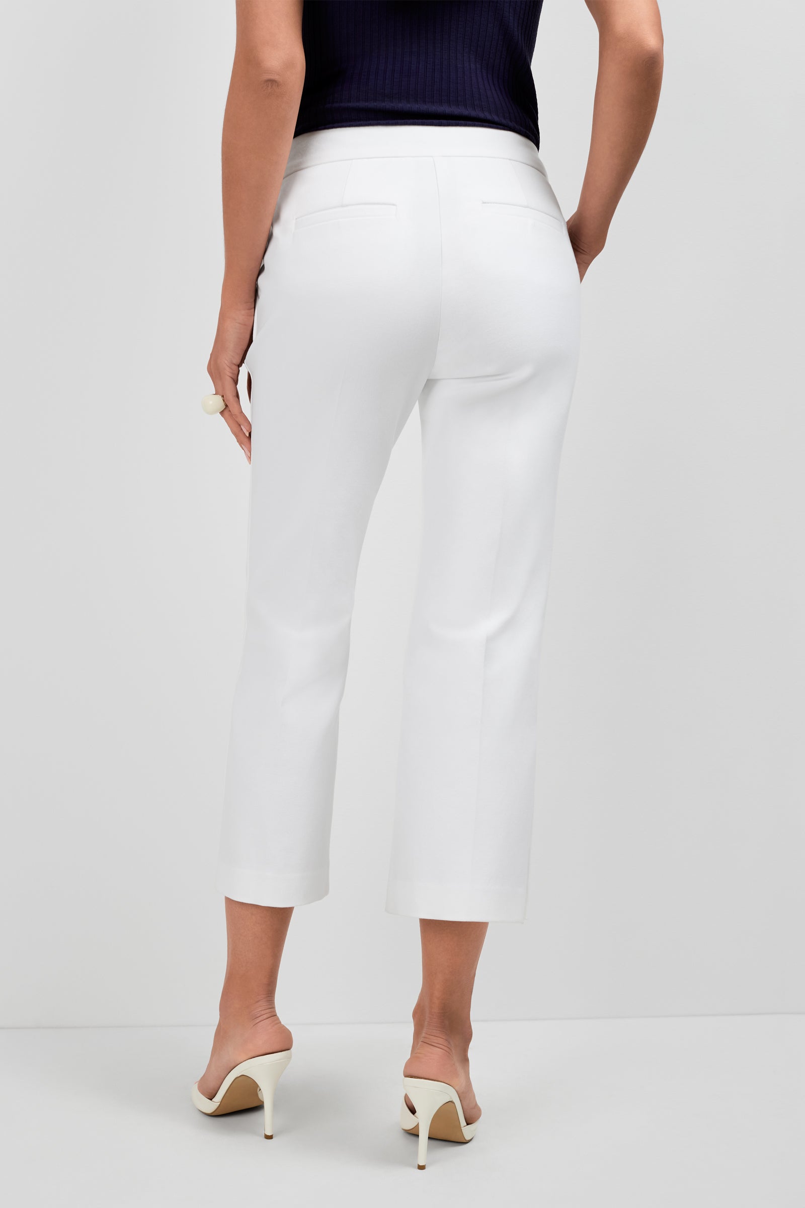 A person is shown from the back wearing Ore Button-Hem Cropped Pants in white, a navy sleeveless top, and white heeled sandals against a plain light gray background.