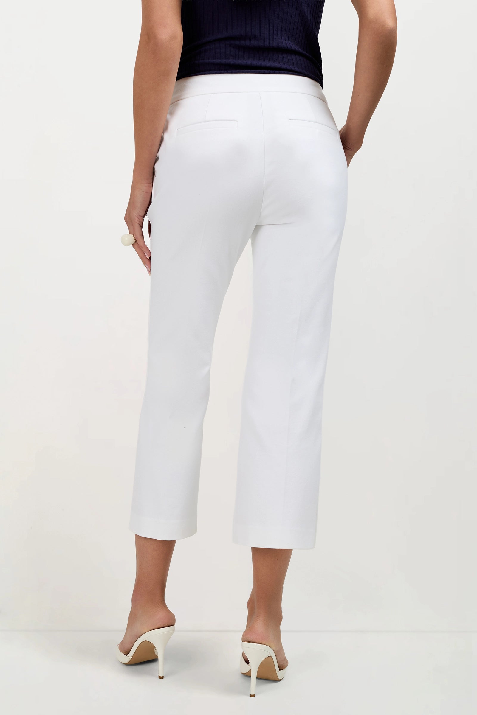 A woman stands facing away, wearing Ore Button-Hem Cropped Pants, a sleeveless black top, and white heeled sandals against a plain white background.