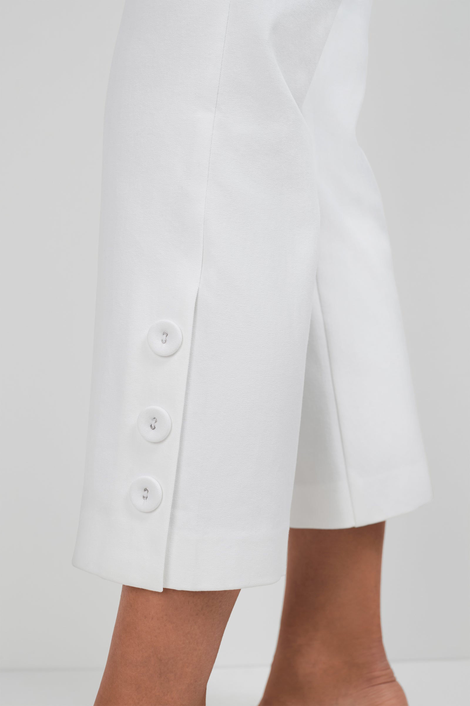 Close-up of a person wearing the Ore Button-Hem Cropped Pants, featuring three white buttons near the outer hem. Only the lower leg and ankle are shown against a plain background.