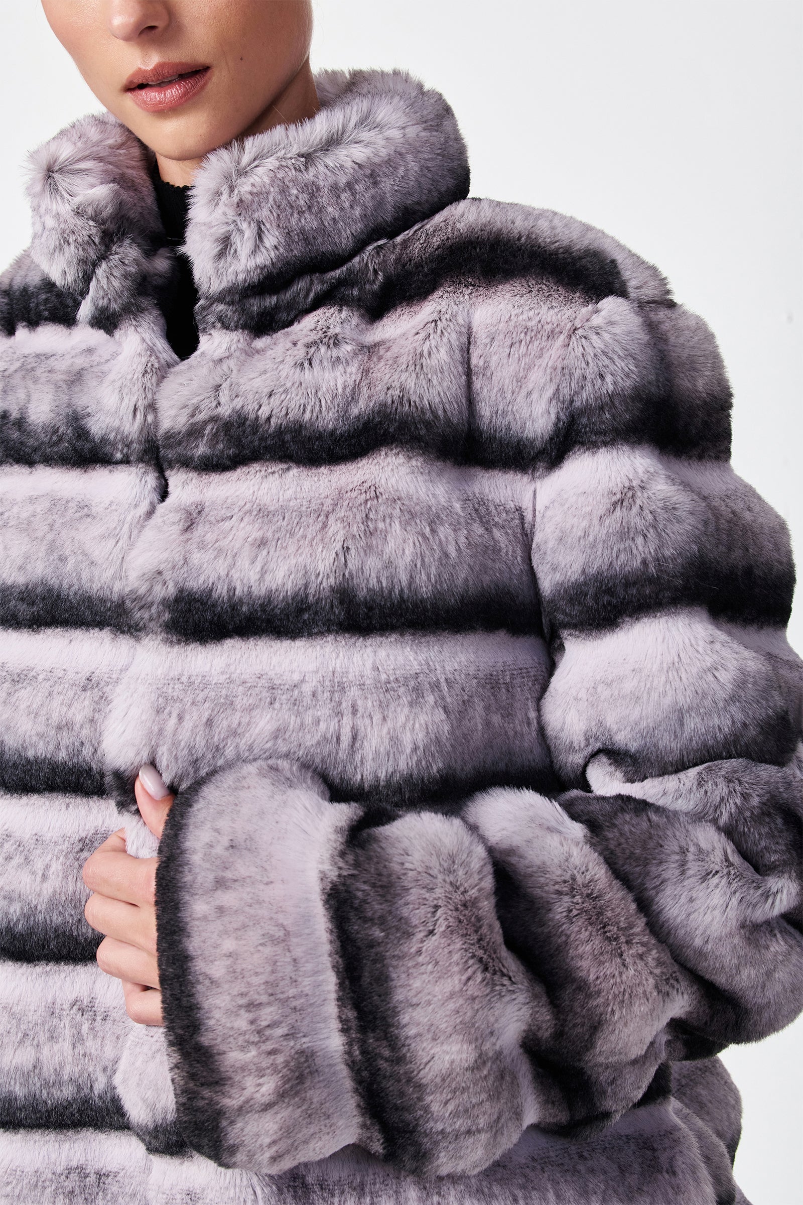A person models the Mikel 2 Faux Fur Mid-Length Coat, a plush striped piece in gray and lavender, with one hand holding the front. The photo is cropped to show only the lower face and upper body.