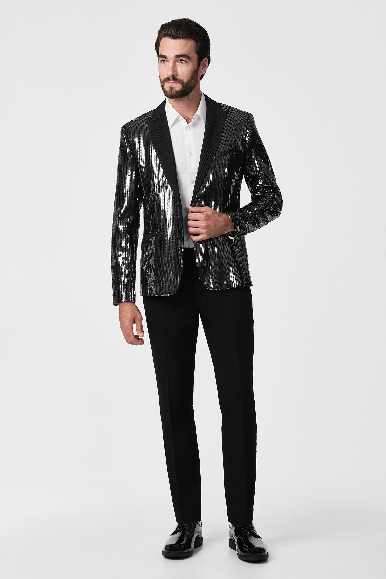 A man stands against a plain background wearing the Adrian Tux Trouser with a shiny black tuxedo jacket featuring vertical stripes, a white dress shirt, and black dress shoes.