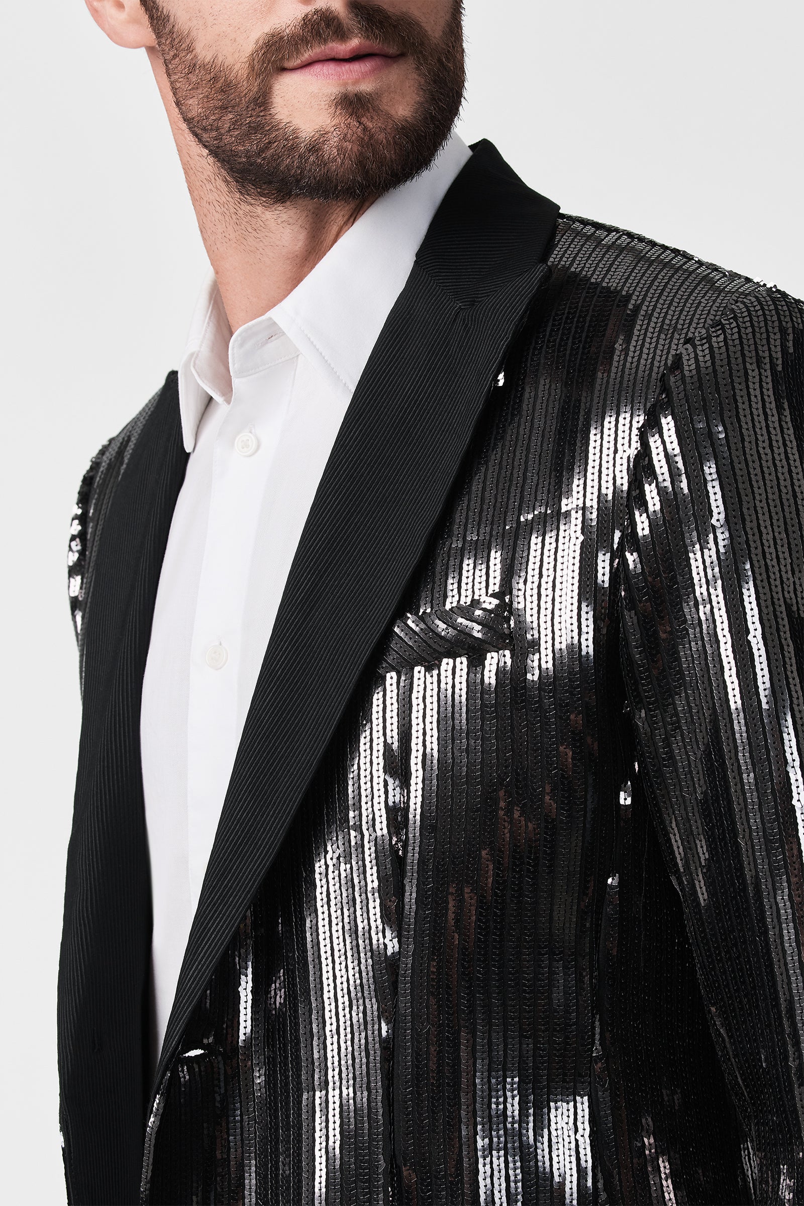 A man with a short beard wears a white dress shirt and the Coleman Blazer, a shiny metallic black sequin jacket with black lapels. The photo is cropped to show part of his face and upper torso.