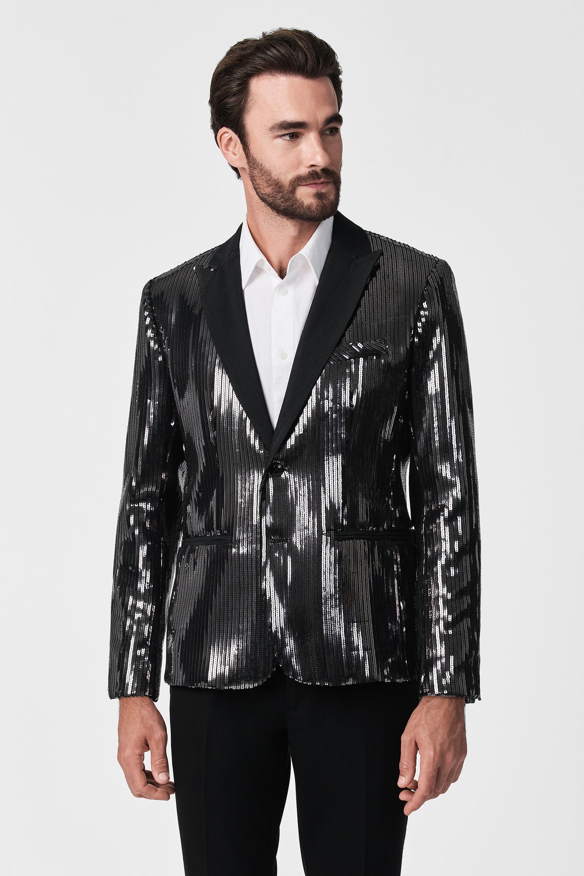 A man with dark hair and a beard models the shiny black Coleman Blazer over a white dress shirt and black pants, standing against a plain white background.