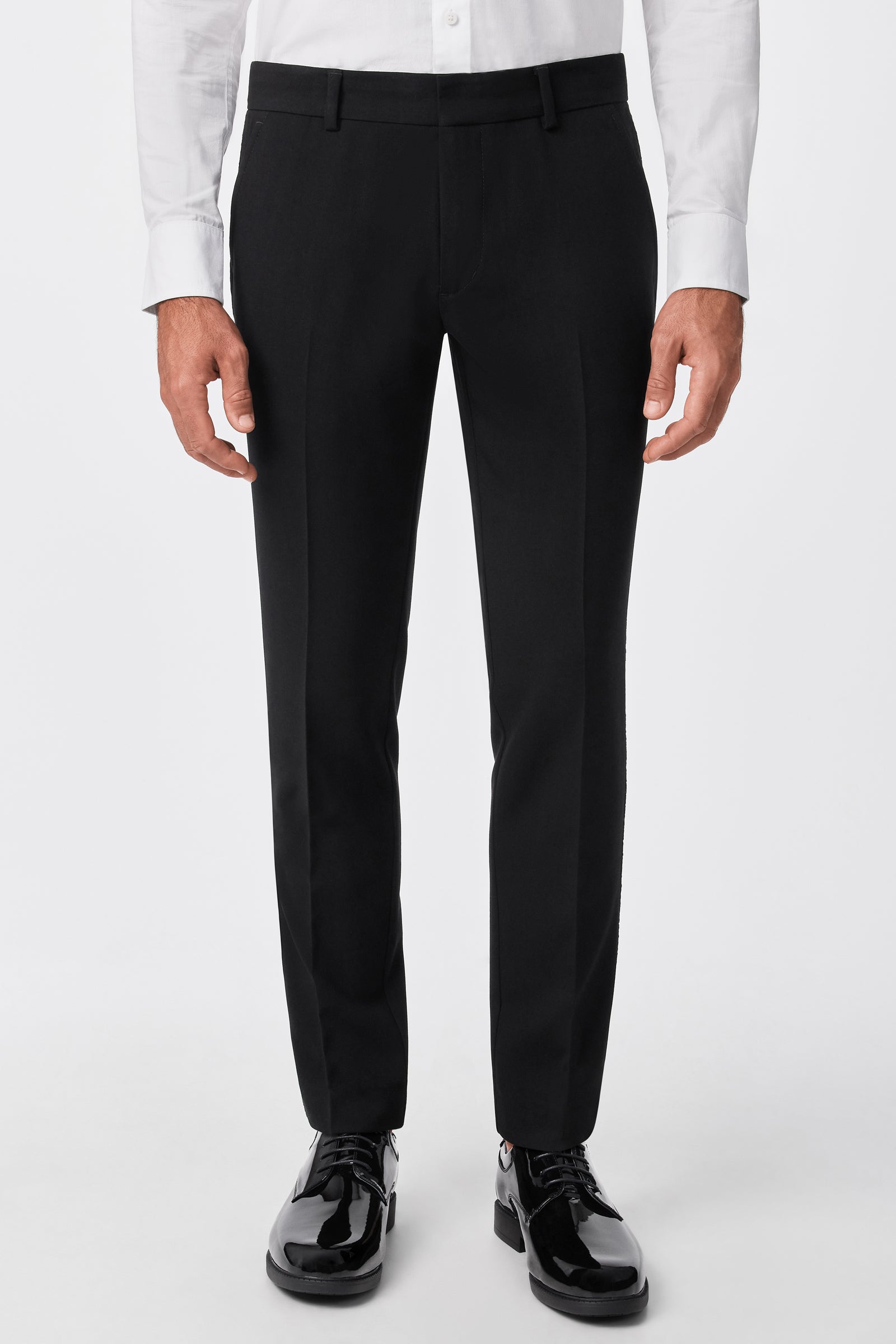 A person wearing the Adrian Tux Trouser in black, paired with a white dress shirt and black shiny dress shoes, stands against a plain white background. The image is cropped from the waist down.