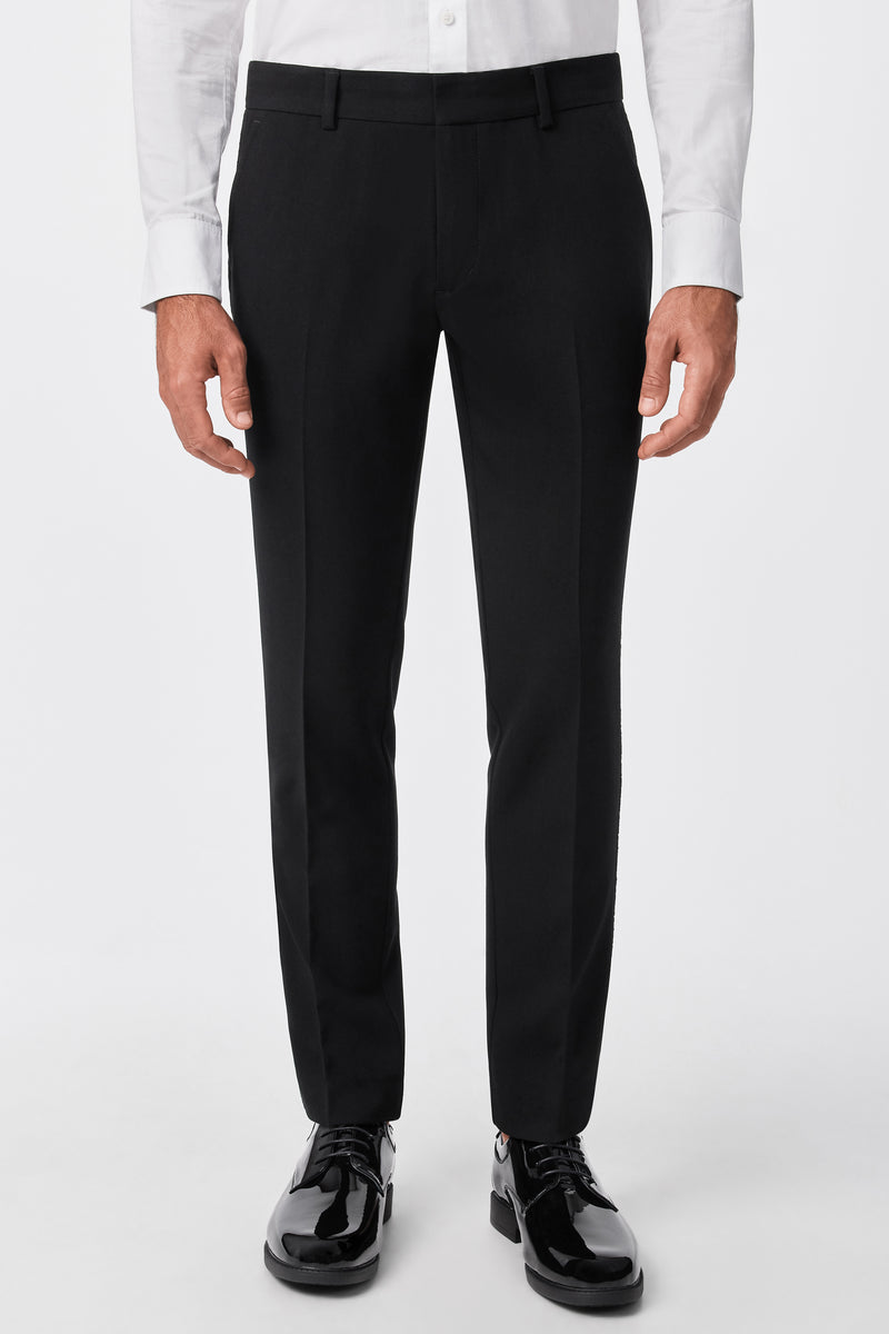 A person wearing the Adrian Tux Trouser in black, paired with a white dress shirt and black shiny dress shoes, stands against a plain white background. The image is cropped from the waist down.