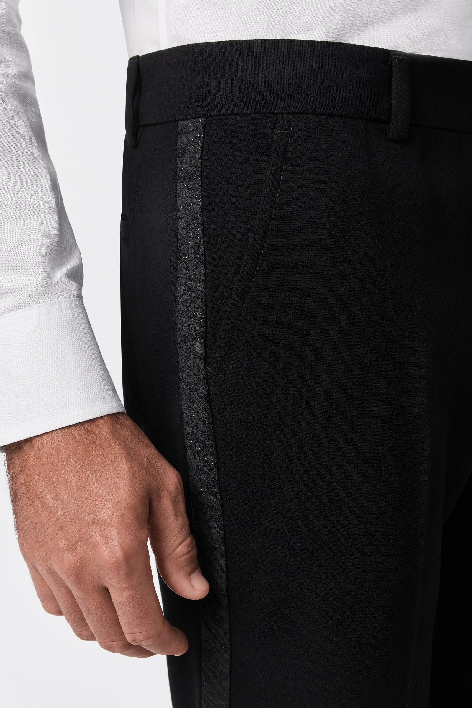 A close-up of a person wearing the Adrian Tux Trouser, black dress pants with a subtle textured side stripe, paired with a white dress shirt; the lower torso and right hand are visible at the side.