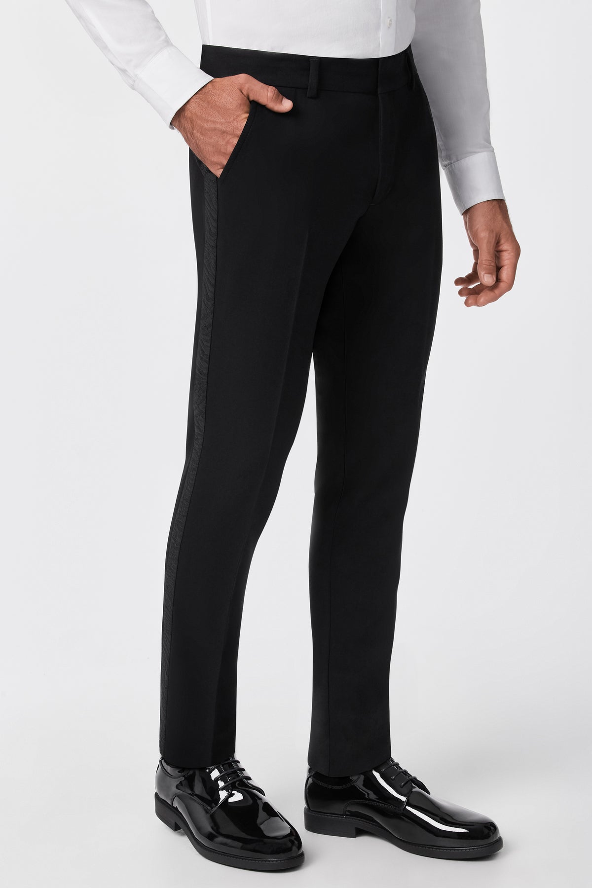A man stands against a plain background with one hand in his pocket, wearing the Adrian Tux Trouser featuring a satin side stripe, paired with a white dress shirt and black shiny dress shoes.