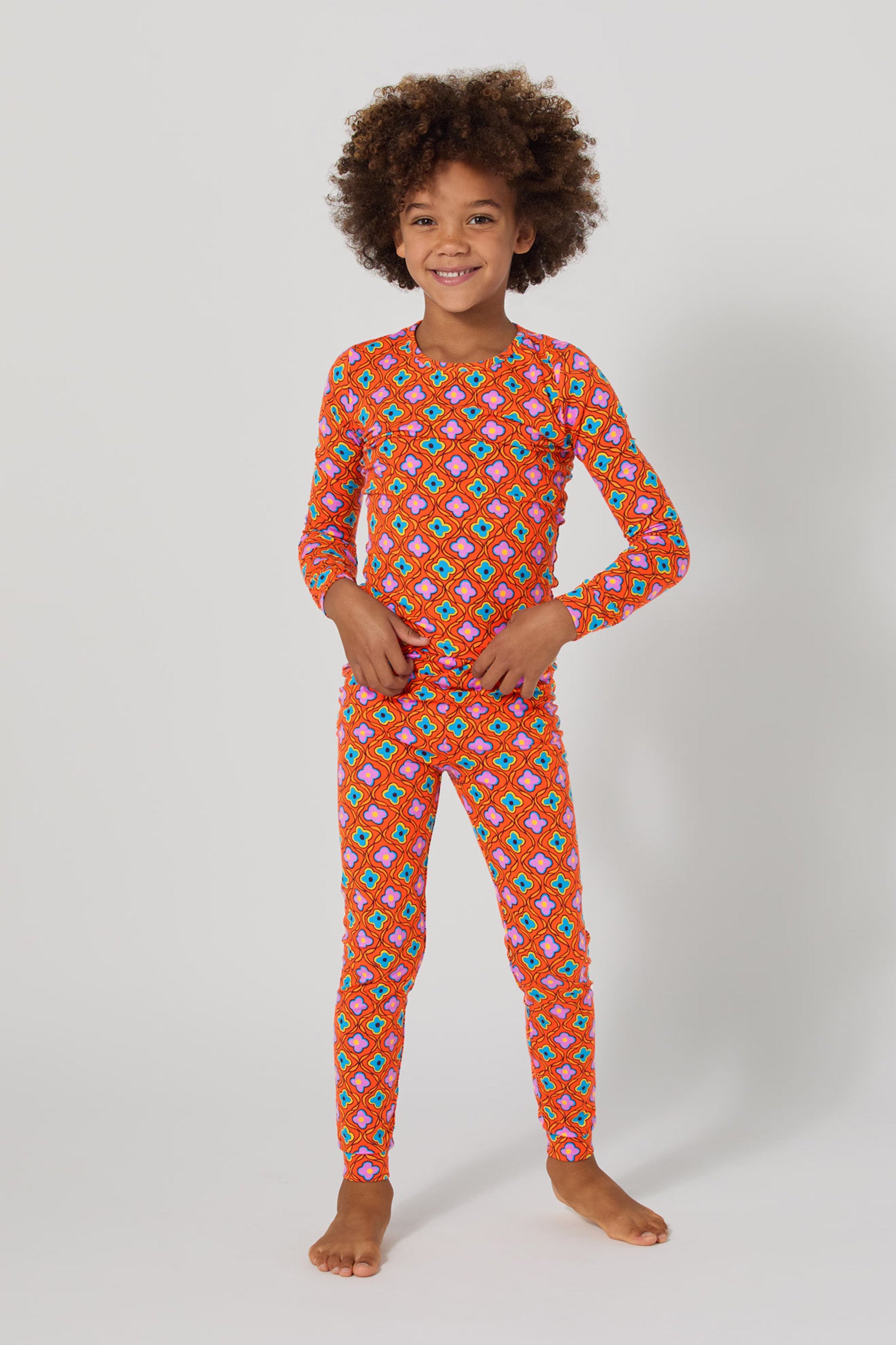 A smiling child with curly hair stands barefoot against a plain background, wearing the Daisy Foulard 2Pc Kids Pj Set—matching long-sleeve top and pants featuring a bright, colorful geometric pattern.