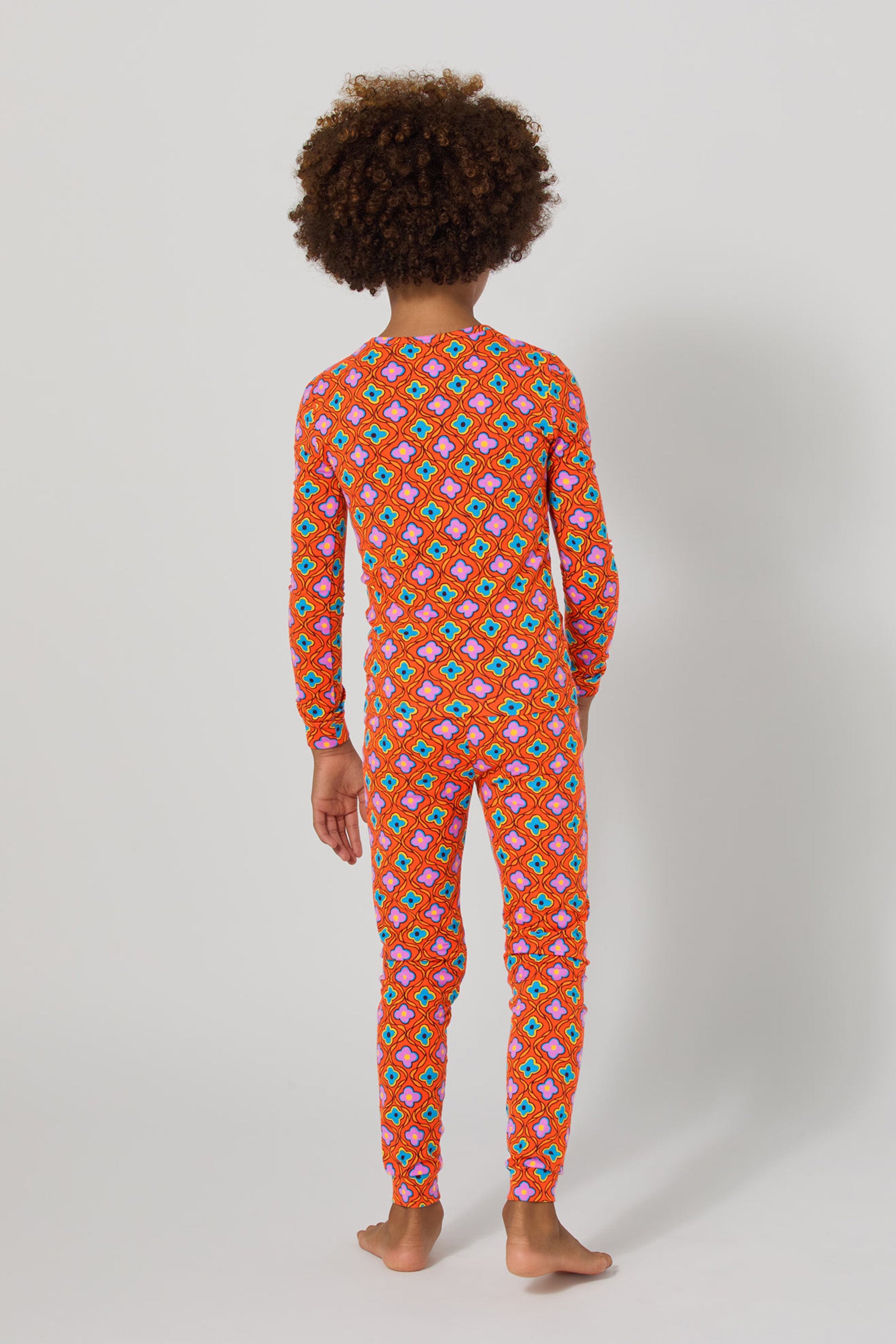 A child with curly hair stands barefoot facing away in the Daisy Foulard 2Pc Kids Pj Set—a red, long-sleeved, full-length pajama set featuring a colorful geometric pattern—against a plain light background.