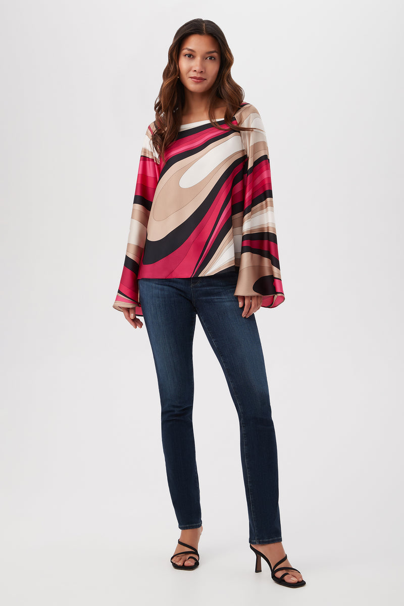 A woman stands against a plain background wearing the Torie Top in Wynwood Waves Satin Georgette—featuring colorful abstract patterns and wide sleeves—paired with dark blue skinny jeans and black heeled sandals.