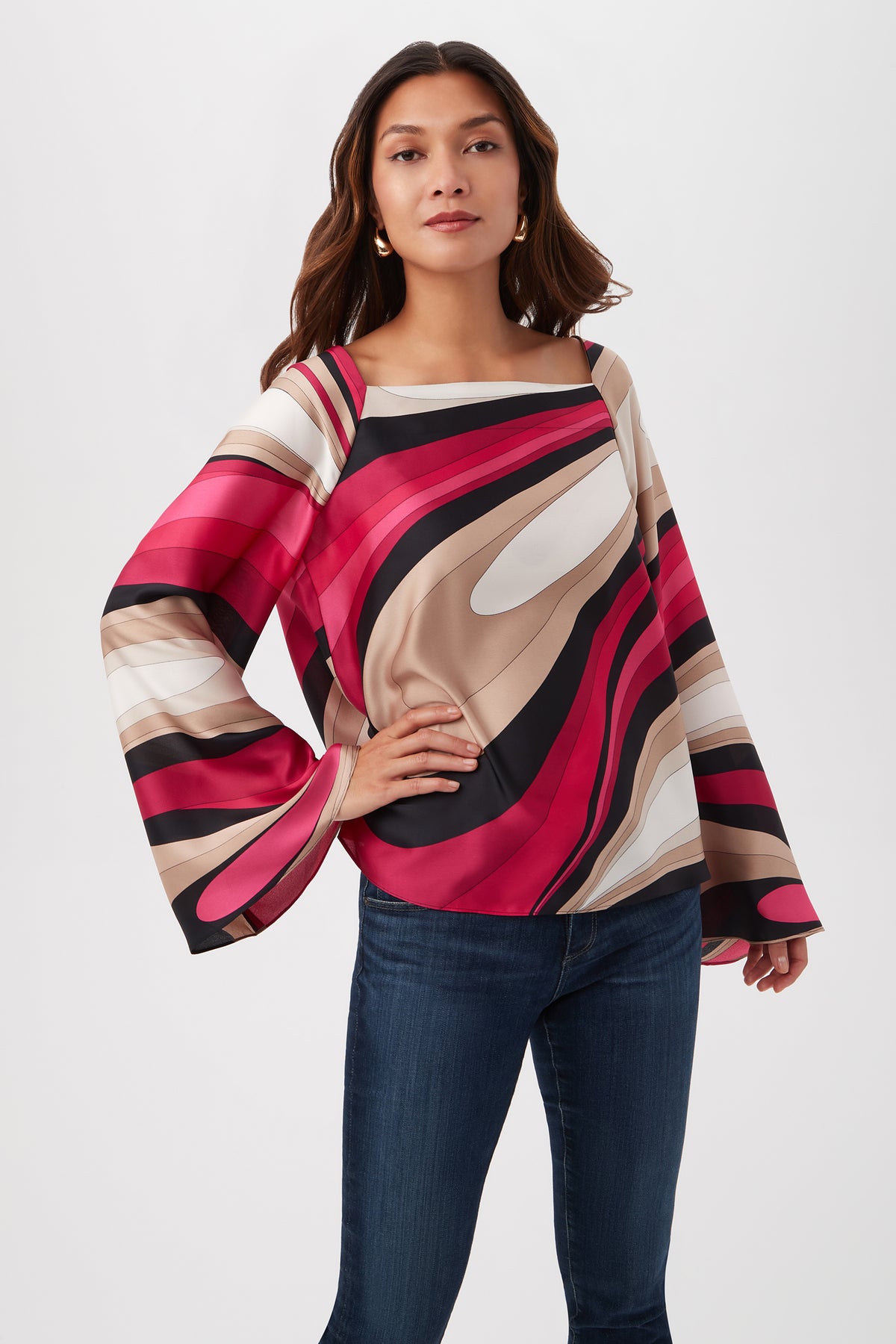 A woman stands confidently in the Torie Top - Wynwood Waves Satin Georgette in Fairy Fuchsia Multi, featuring bold wavy patterns and long sleeves, paired with dark blue jeans against a white background.