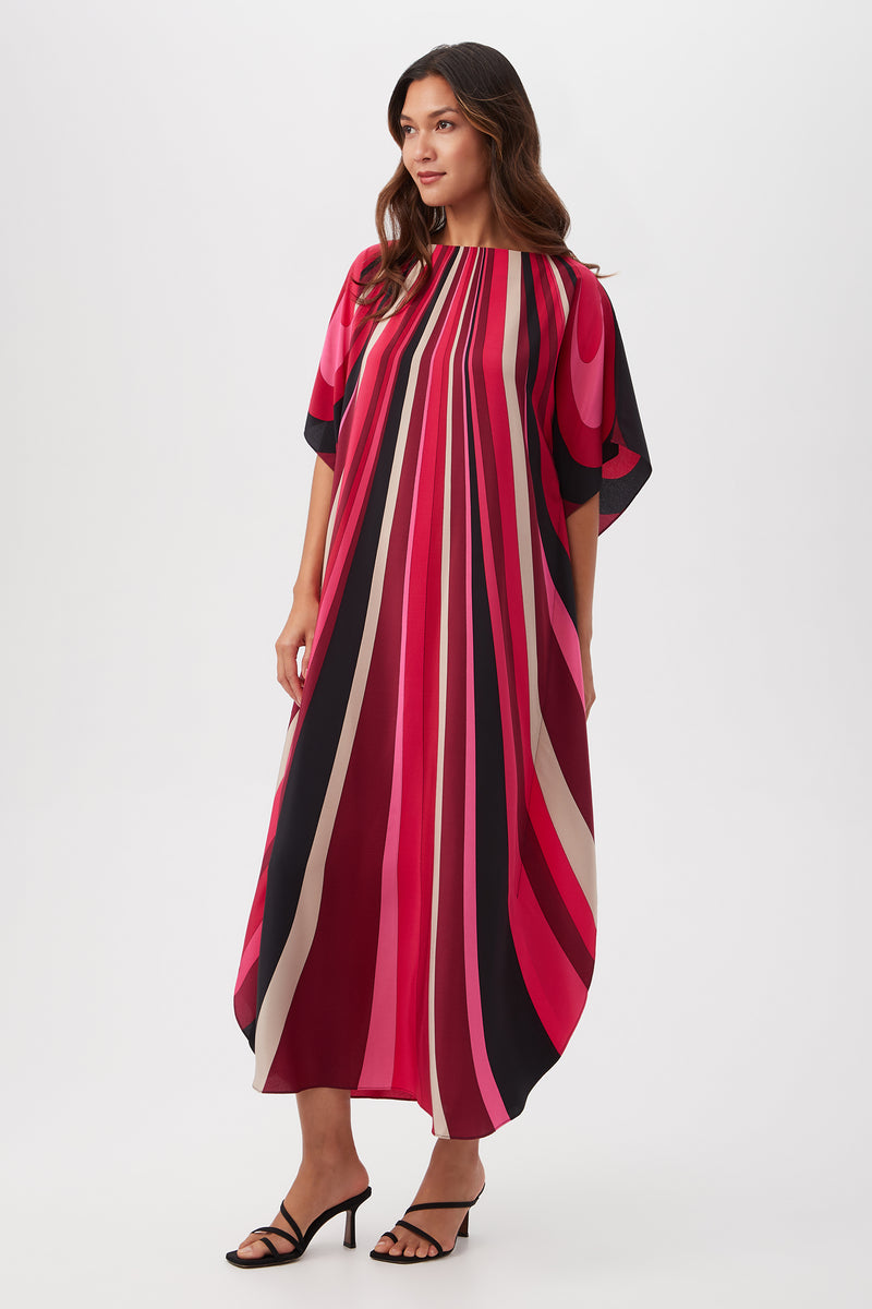 A woman wears the Jalani Maxi Caftan Dress - Wynwood Waves Silk, a loose, ankle-length dress with bold vertical pink, red, black, and beige stripes. It features wide short sleeves and a round neckline. She pairs it with black heeled sandals against a white background.