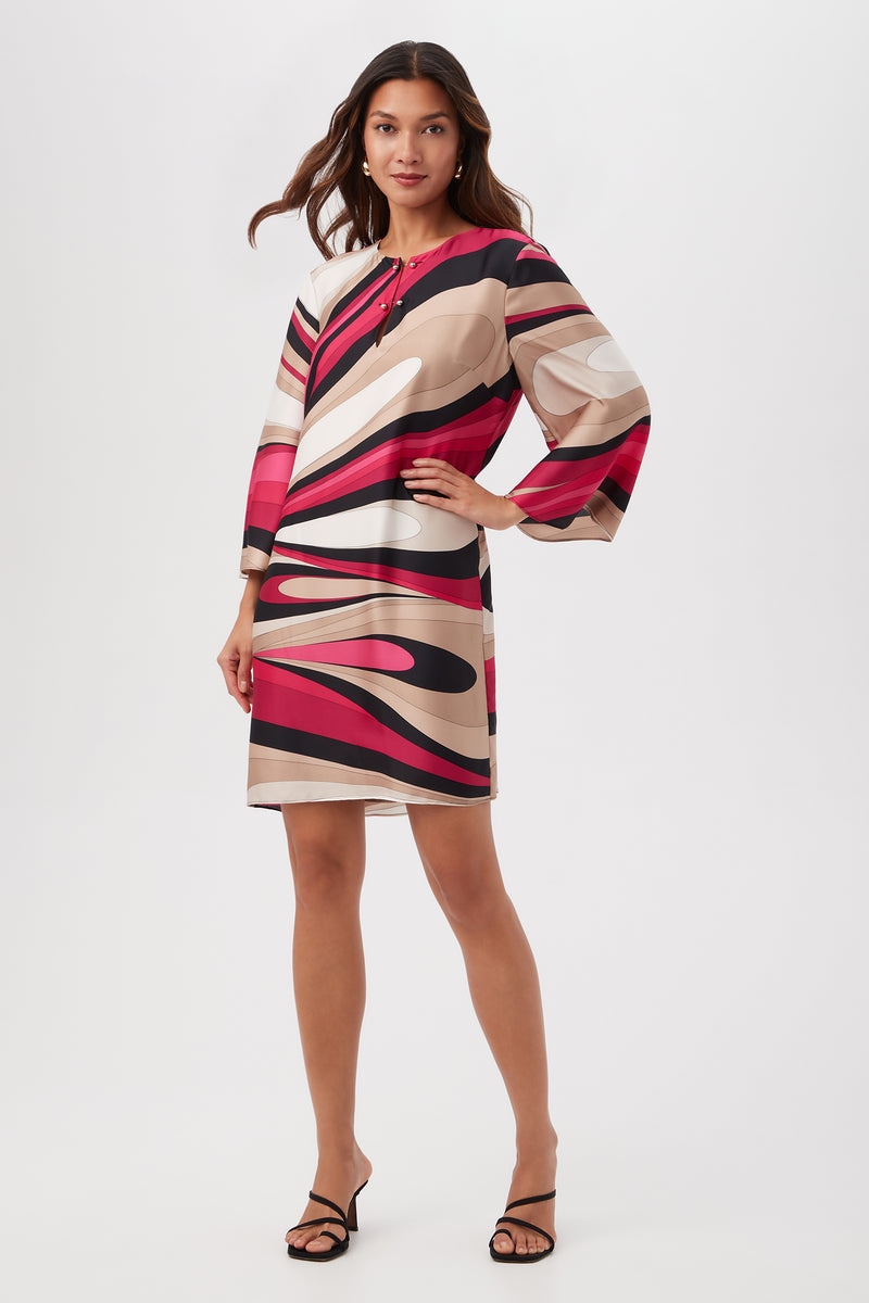 A woman poses with one hand on her hip in the Nallia Dress - Wynwood Waves Satin Georgette, featuring a Fairy Fuchsia Multi abstract pattern. She has long wavy hair and wears black sandals against a white background.