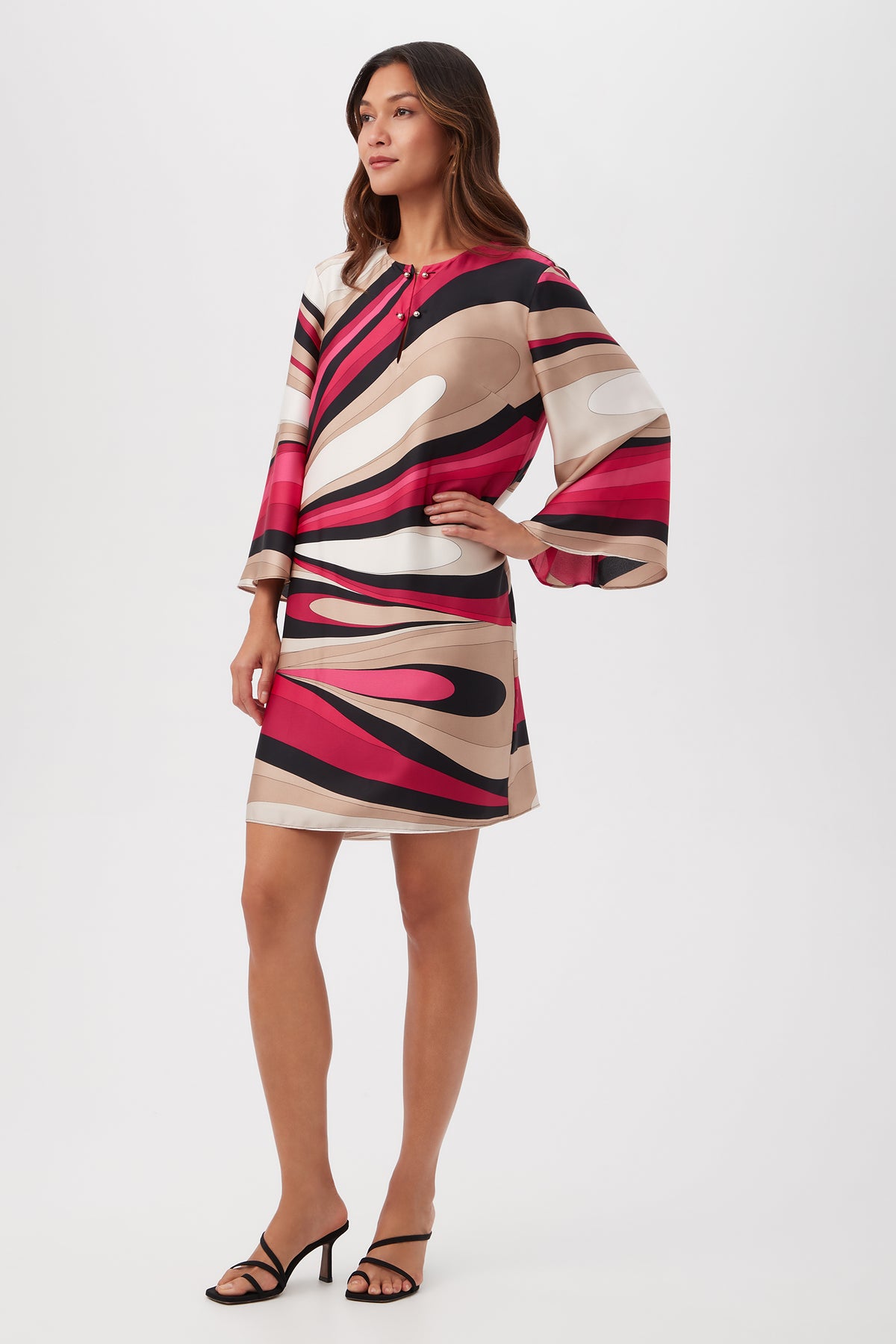 A woman wears the Nallia Dress - Wynwood Waves Satin Georgette, featuring a colorful abstract pattern, long sleeves, and a short hemline. She pairs it with black heeled sandals against a plain white background.