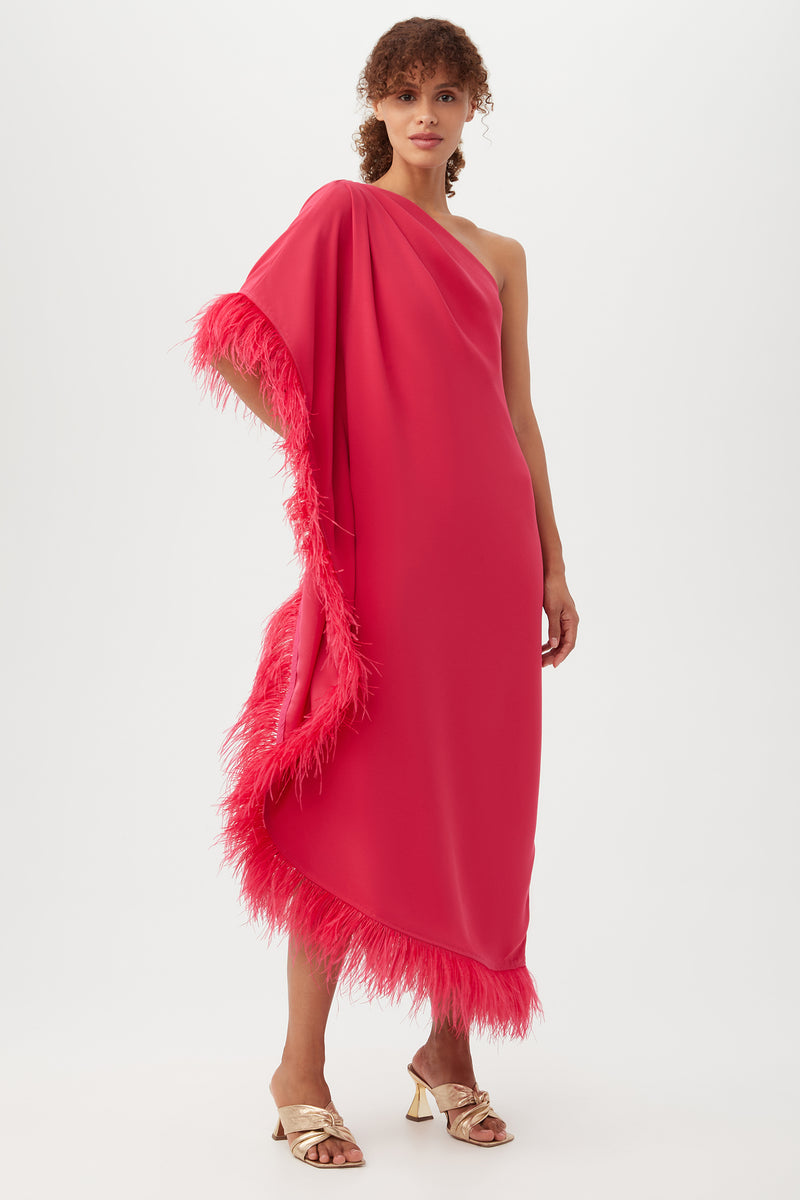 A woman models the Largo Caftan Dress, a vibrant pink one-shoulder style with feather trim, paired with gold heeled sandals, against a plain white background.