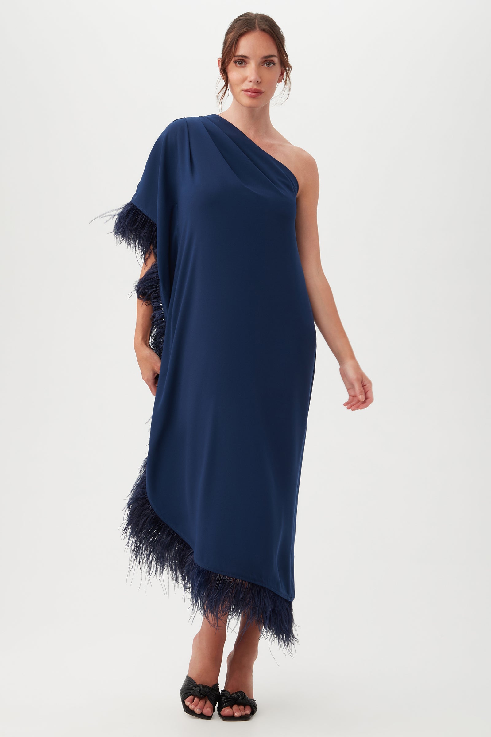 A woman wears the Largo Caftan Dress, a navy one-shoulder style featuring feather trim, standing against a plain white background.