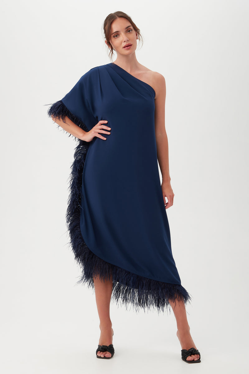 A woman stands against a plain white background wearing the Largo Caftan Dress in Ink, featuring a one-shoulder design with feather trim along the asymmetric hem and side seam. Her hair is tied back and she pairs the dress with black open-toe heels.