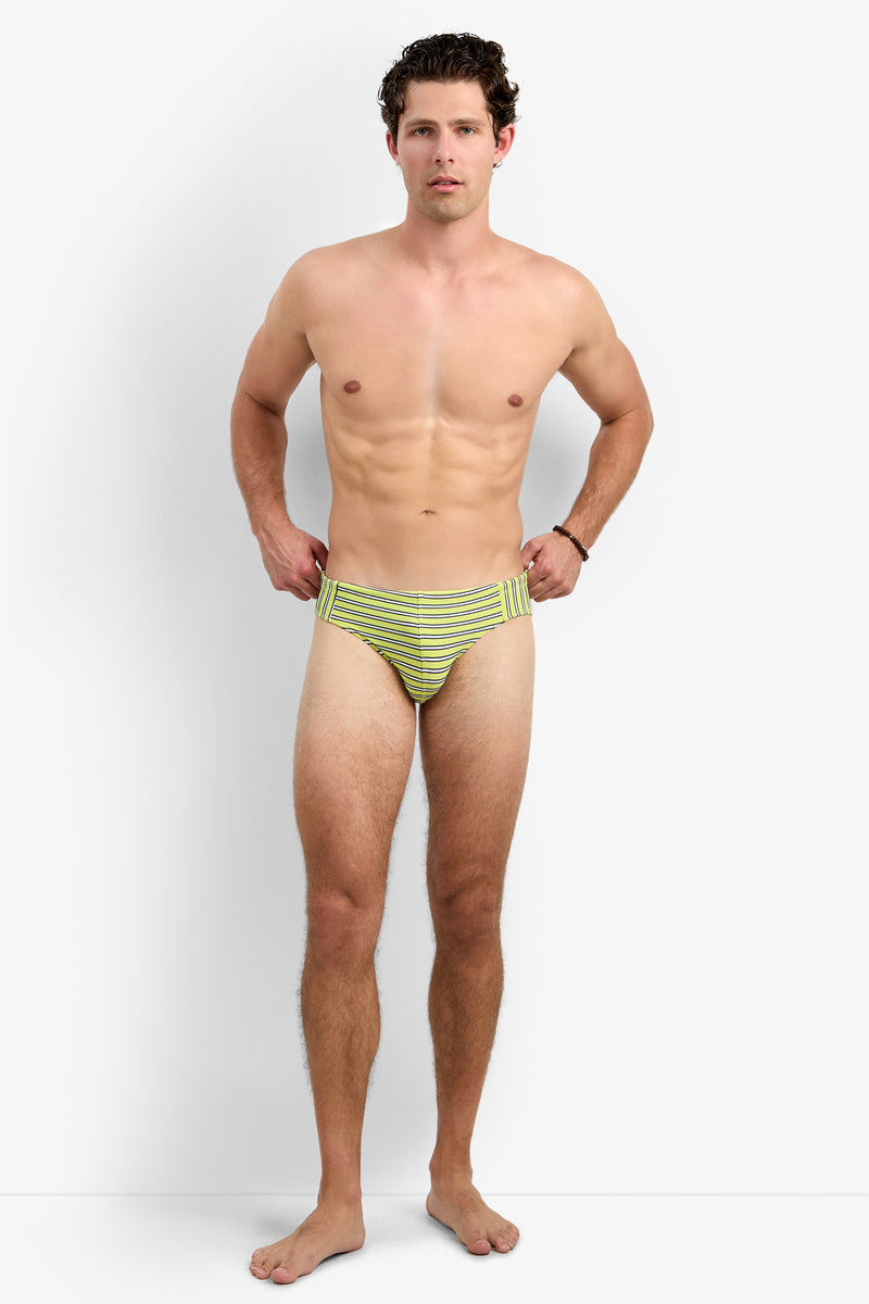 A man stands barefoot against a plain white background, wearing the Gold Coast Swim Brief - Dana Point Stripe in yellow and green stripes, looking forward with his hands on his hips.