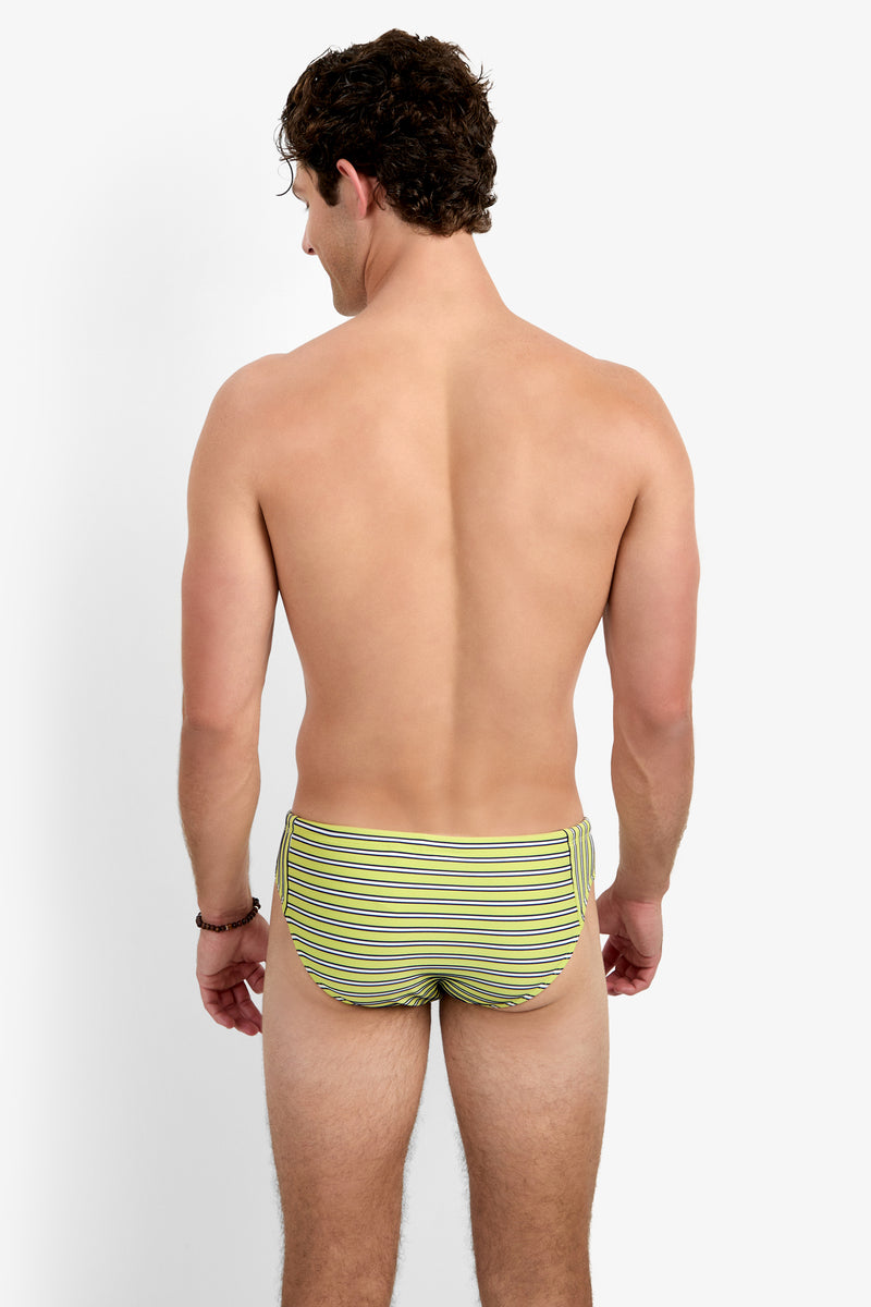 A man with short curly hair faces away from the camera, wearing the Gold Coast Swim Brief - Dana Point Stripe in green, yellow, and white stripes. The background is plain white.