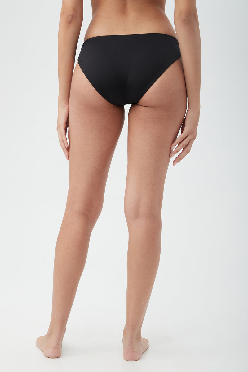A woman stands barefoot against a plain light background, shown from the back wearing the Women's Monaco Shirred Hipster Swim Bottom in black, with her arms resting naturally at her sides.
