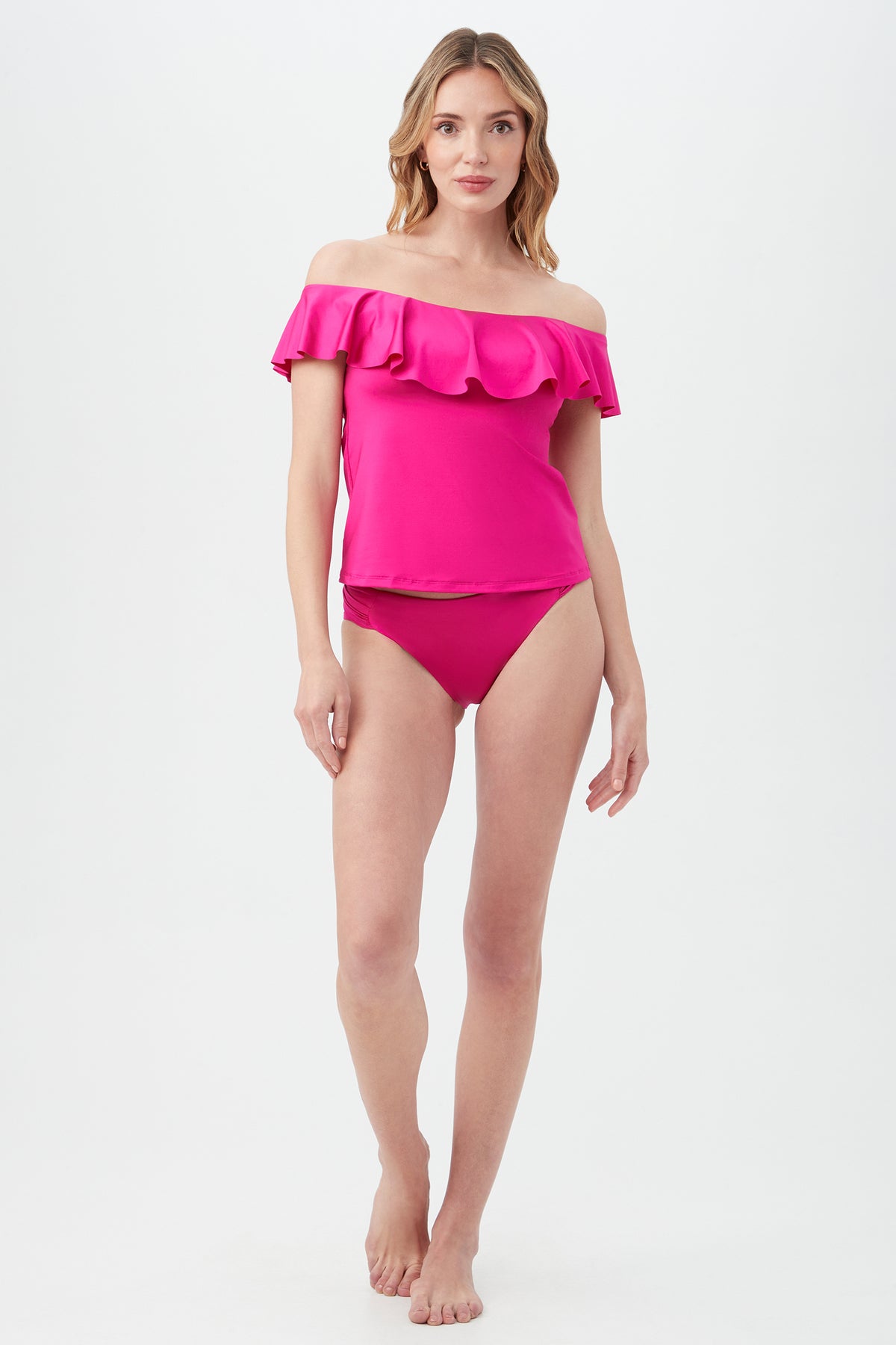 A woman stands barefoot against a white background wearing the Women's Monaco Off The Shoulder Ruffle Tankini Swim Top in bright pink with matching bikini bottoms. She has long, wavy hair and a neutral expression.