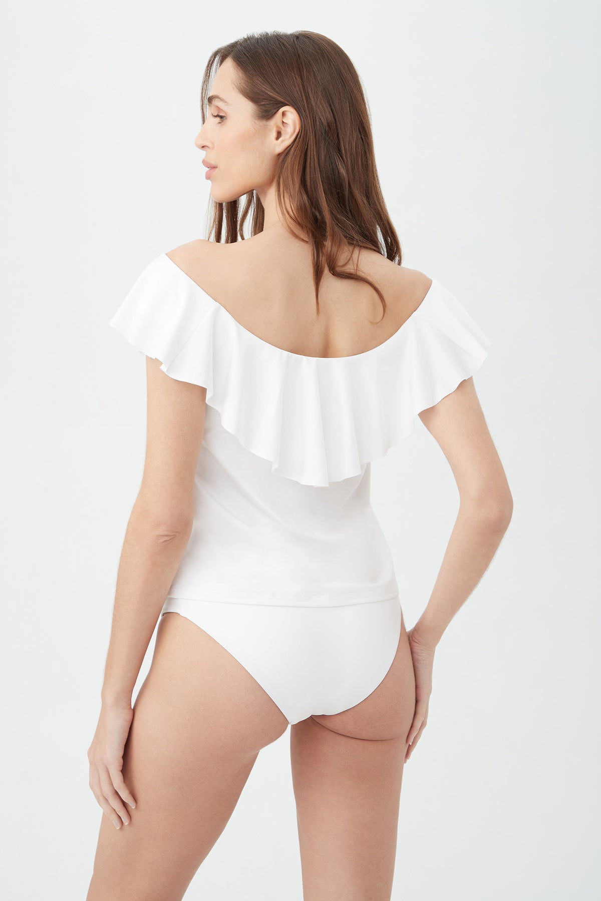 A woman with long brown hair models the Women's Monaco Off The Shoulder Ruffle Tankini Swim Top in white, shown from the back against a plain light background.