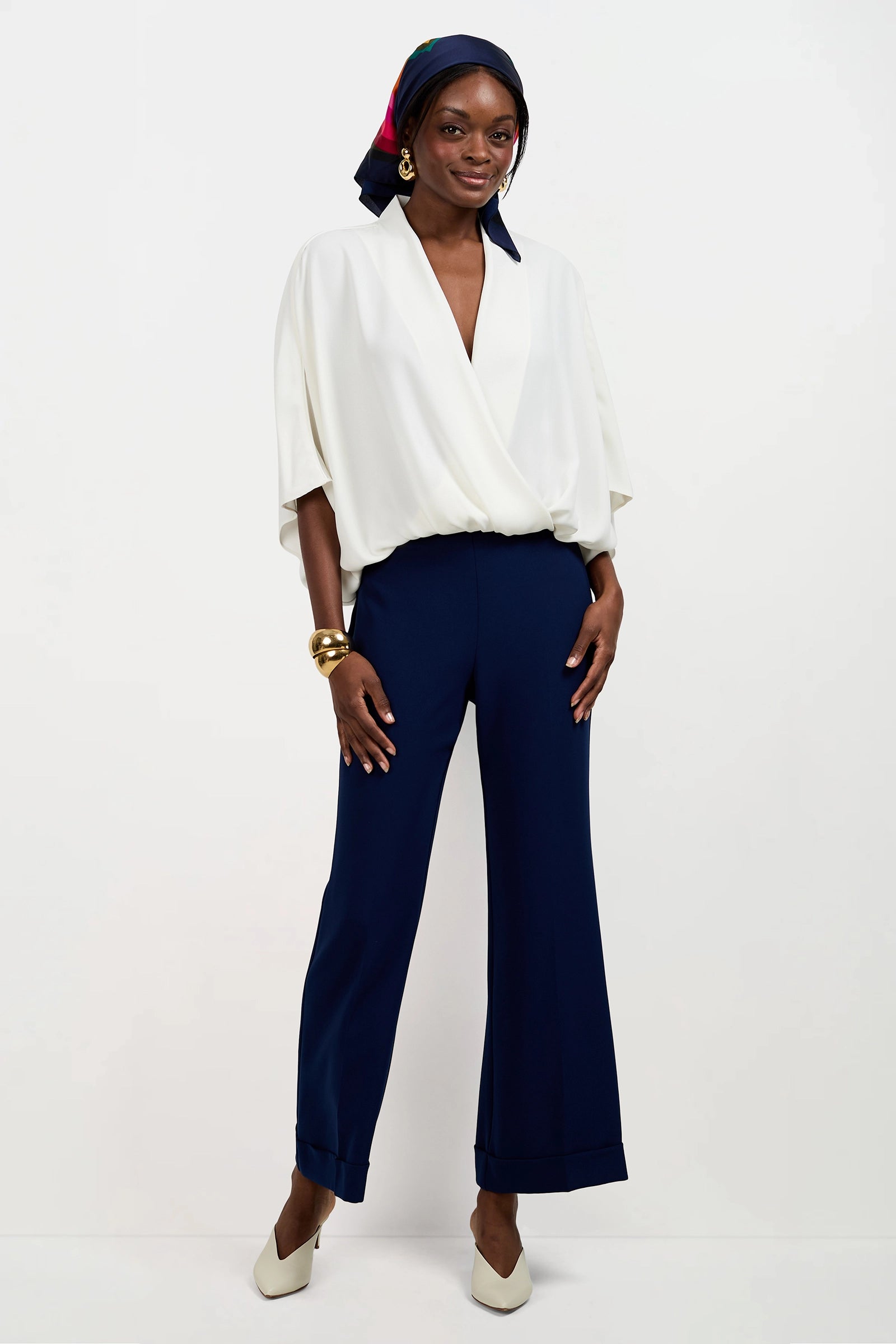 A woman stands against a plain background wearing the Anora V-Neck Wrap Top, navy wide-leg pants, white heels, gold earrings, bangles, and a colorful headscarf tied around her head.