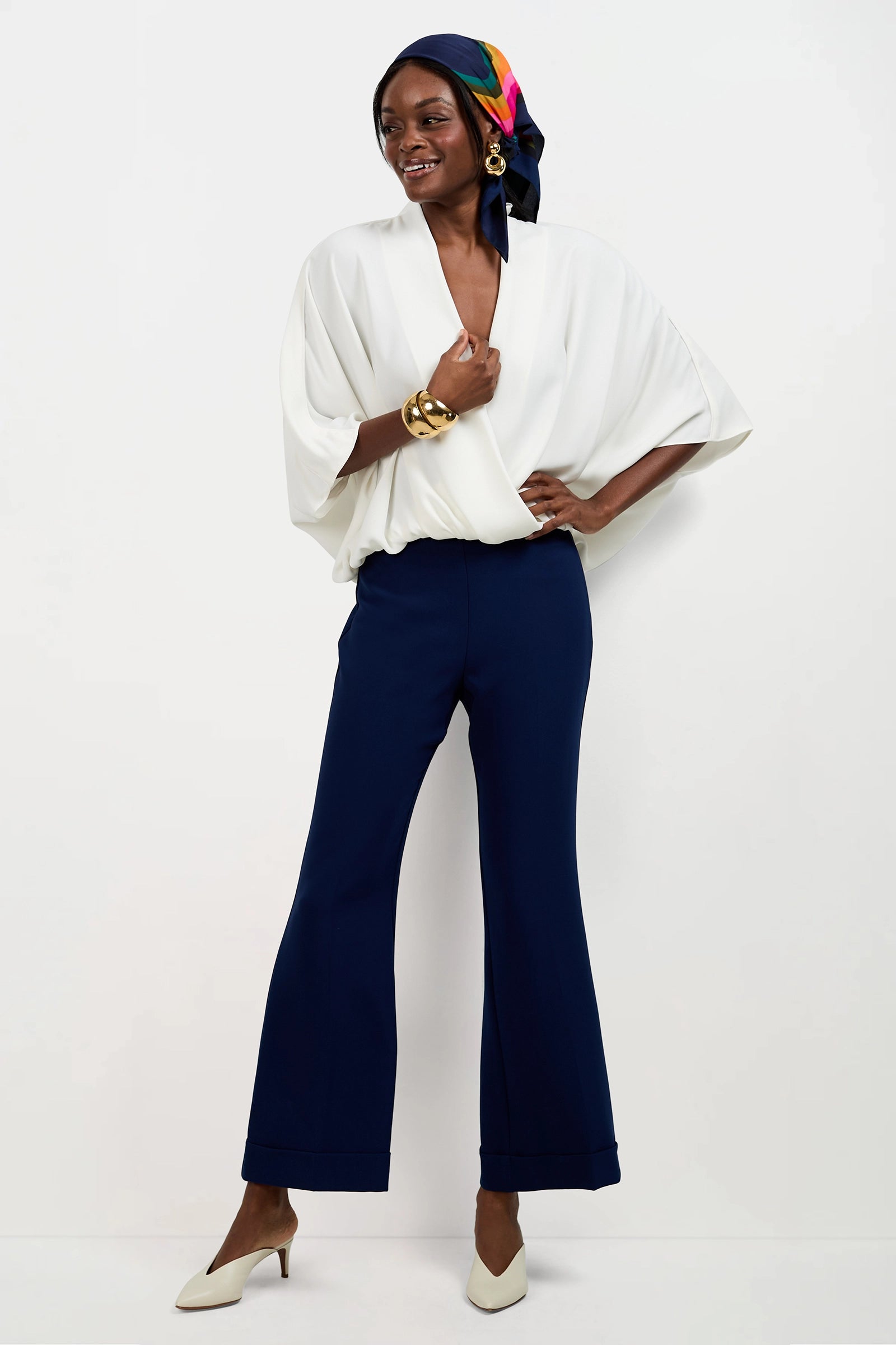 A woman stands against a white background wearing the Anora V-Neck Wrap Top with wide sleeves, navy wide-leg pants, white heels, gold bracelets, large earrings, and a colorful scarf tied around her head.