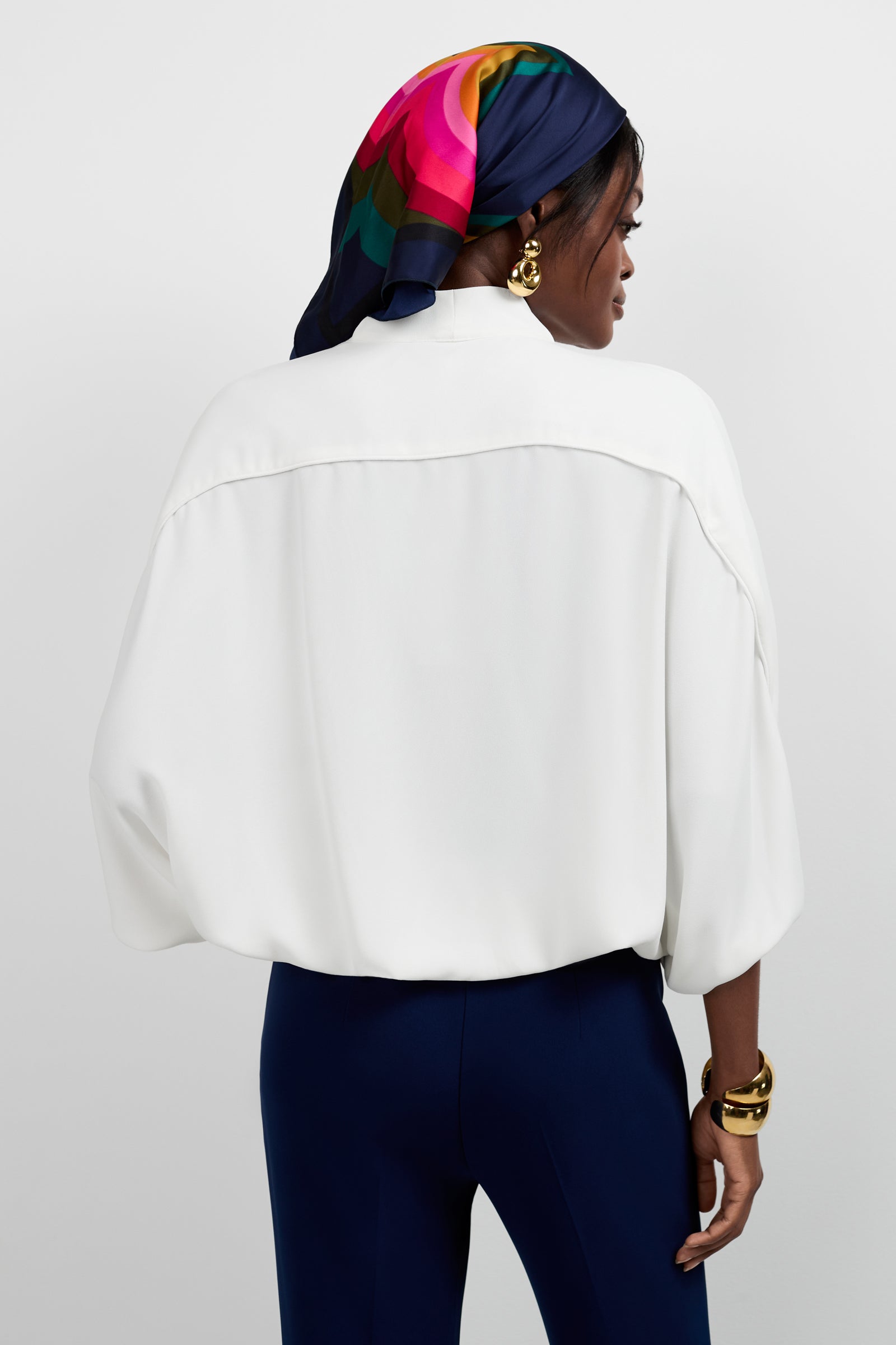 A woman stands facing away, wearing the Anora V-Neck Wrap Top in white, navy pants, and a colorful scarf with pink, red, green, and blue over her hair. She accessorizes with gold hoop earrings and a chunky gold bracelet.