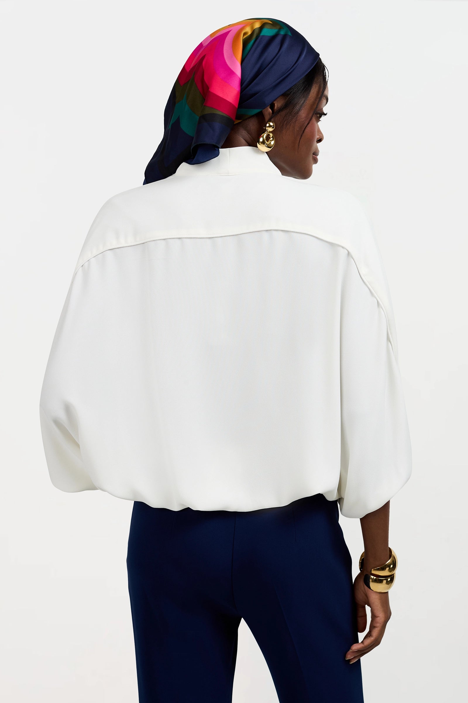 A woman stands facing away, wearing the Anora V-Neck Wrap Top in white, navy pants, gold jewelry, and a colorful silk scarf with pink, green, and blue patterns wrapped over her hair.