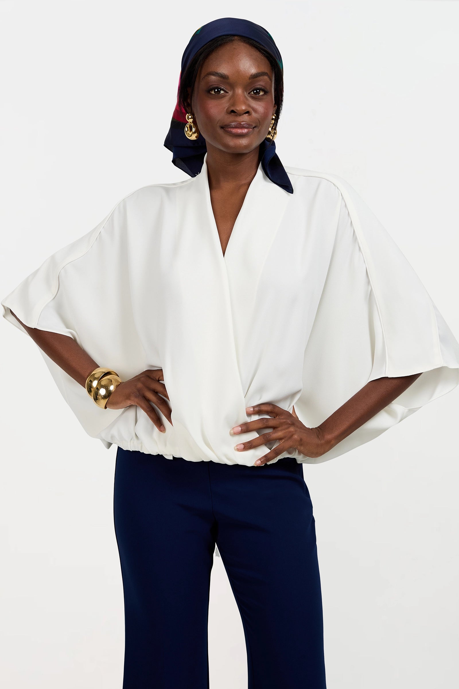 A woman stands confidently with hands on hips, wearing the Anora V-Neck Wrap Top in white, navy pants, a navy and red headscarf, gold hoop earrings, and a chunky gold bracelet against a plain white background.