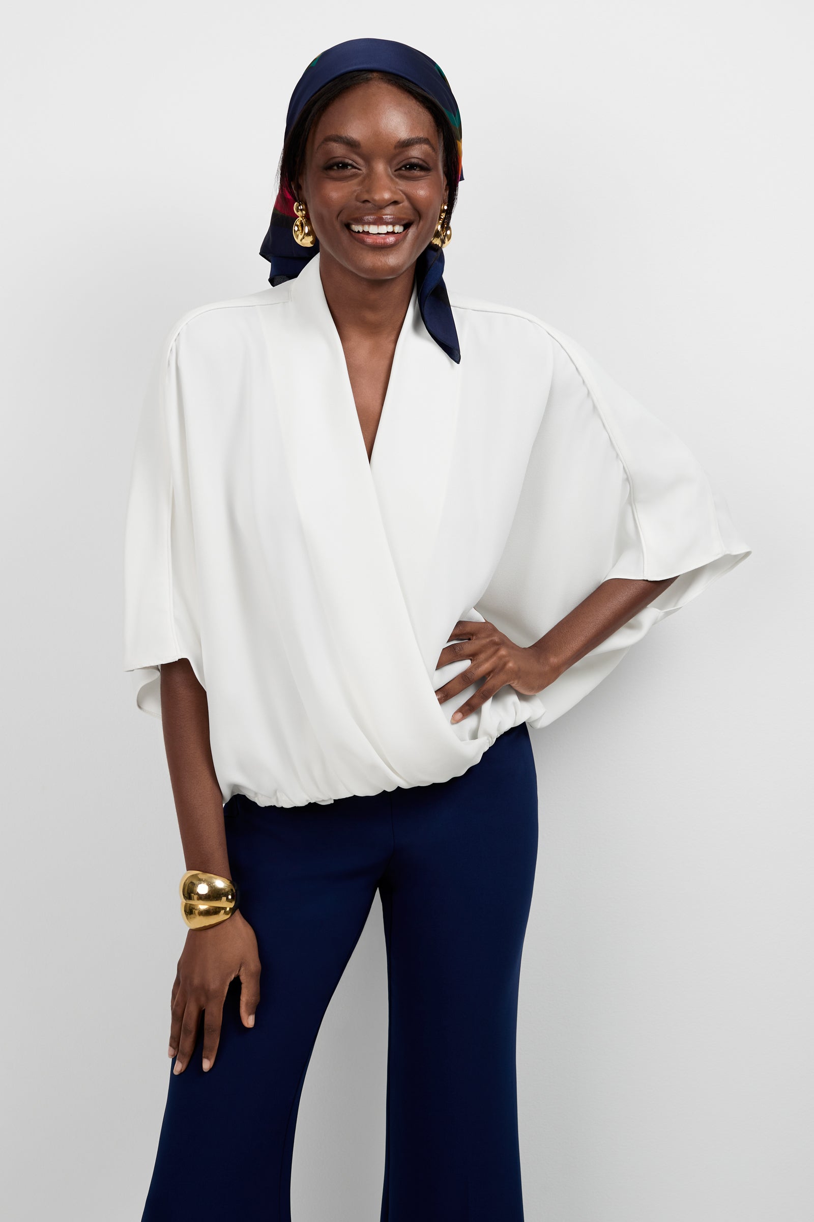 A woman smiles against a white background, wearing the Anora V-Neck Wrap Top in Whitewash, dark blue pants, a colorful headscarf, gold hoop earrings, and a chunky gold bracelet. One hand is resting on her hip.