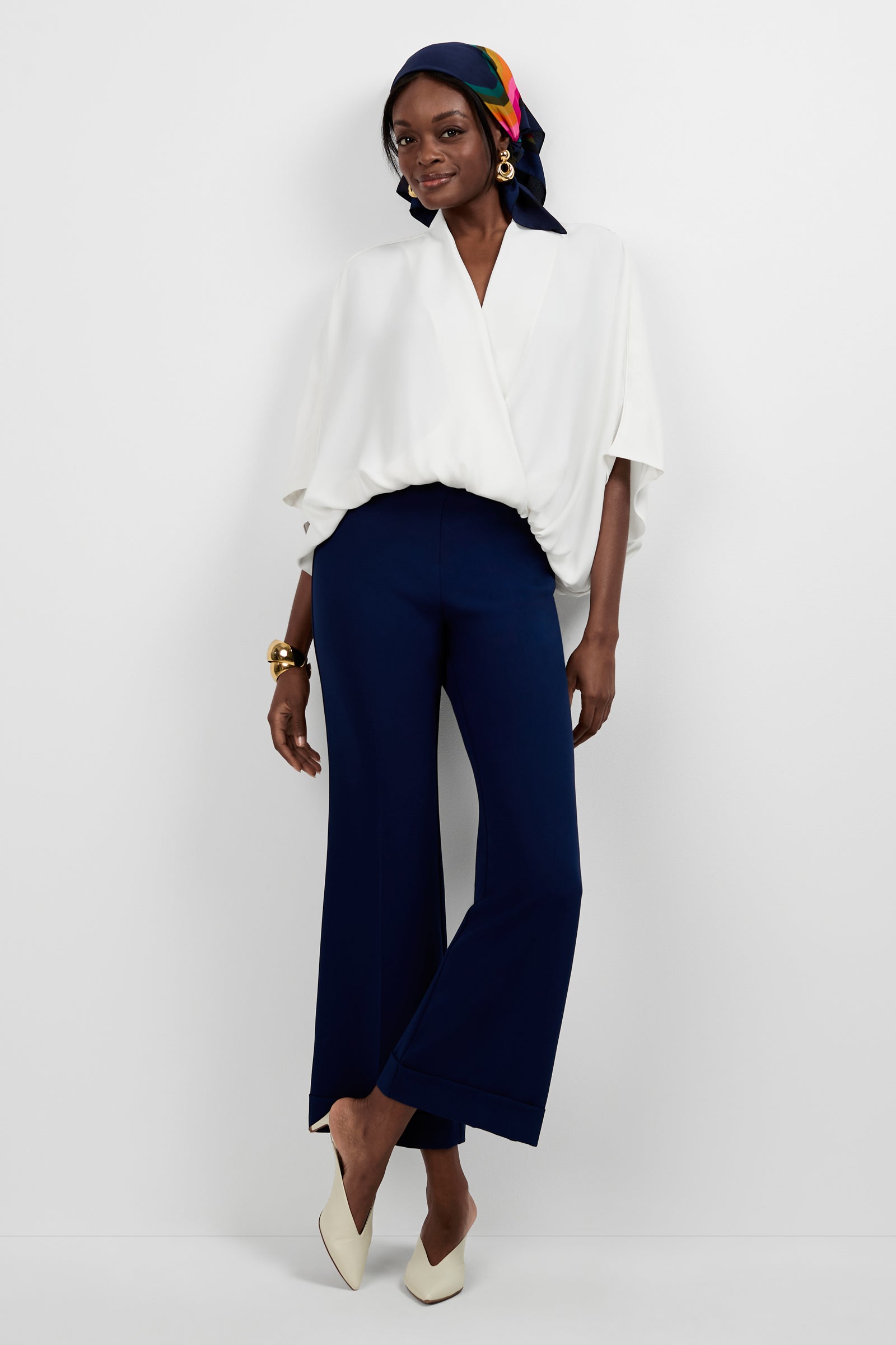 A woman smiles with one arm relaxed at her side, wearing the Anora V-Neck Wrap Top, navy wide-leg pants, white heels, a colorful headscarf, gold earrings, and a chunky gold bracelet against a white background.