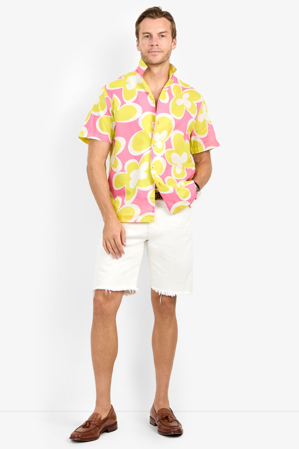 A man wears the Felix Camp Shirt in pink and yellow floral with white frayed shorts, a brown belt, and brown loafers, standing against a plain white background.